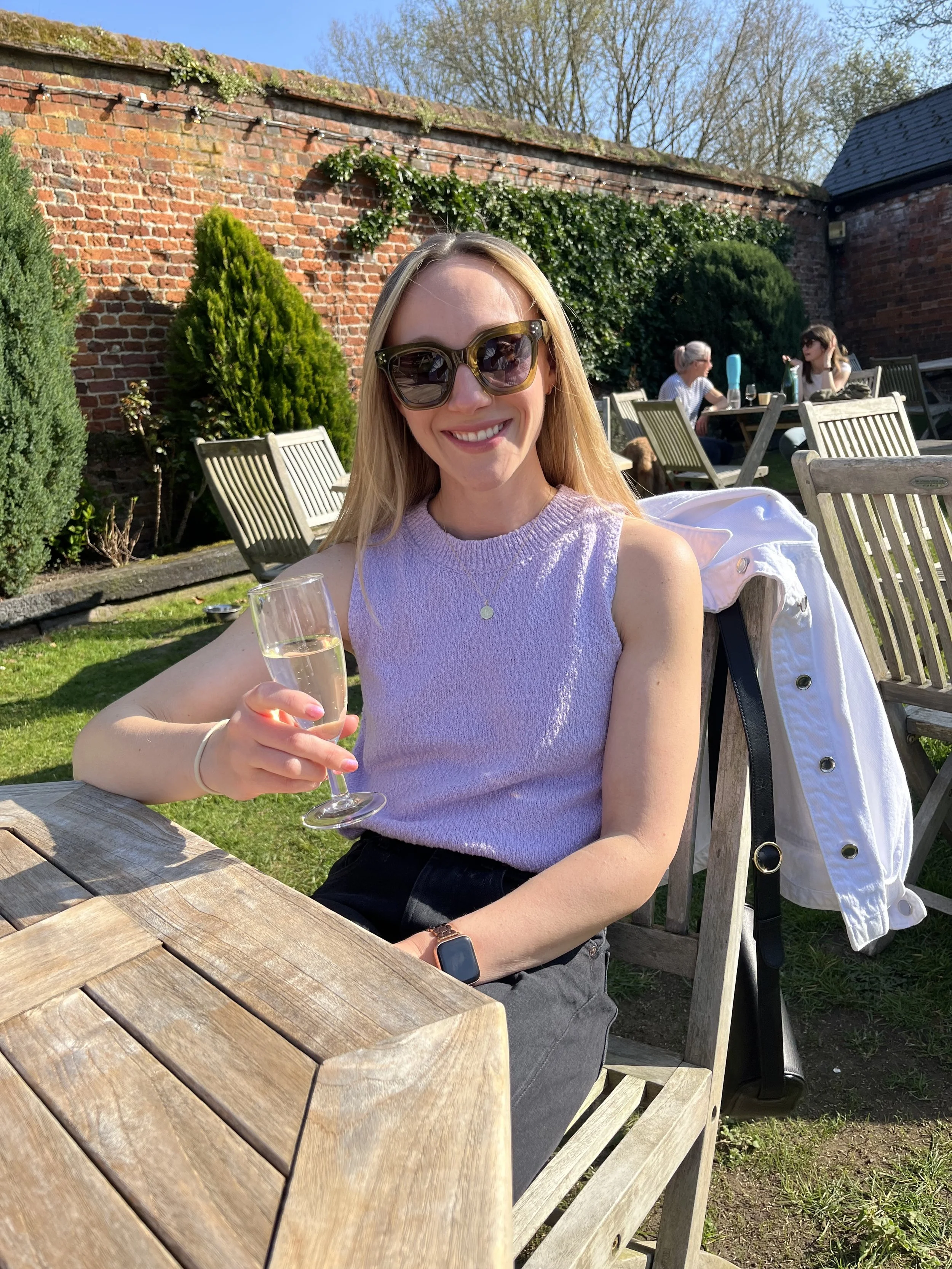 A young woman sitting outdoors at a wooden table, wearing sunglasses and a sleeveless lavender top, holding a glass of champagne, smiling, with other people and a brick wall with greenery in the background.