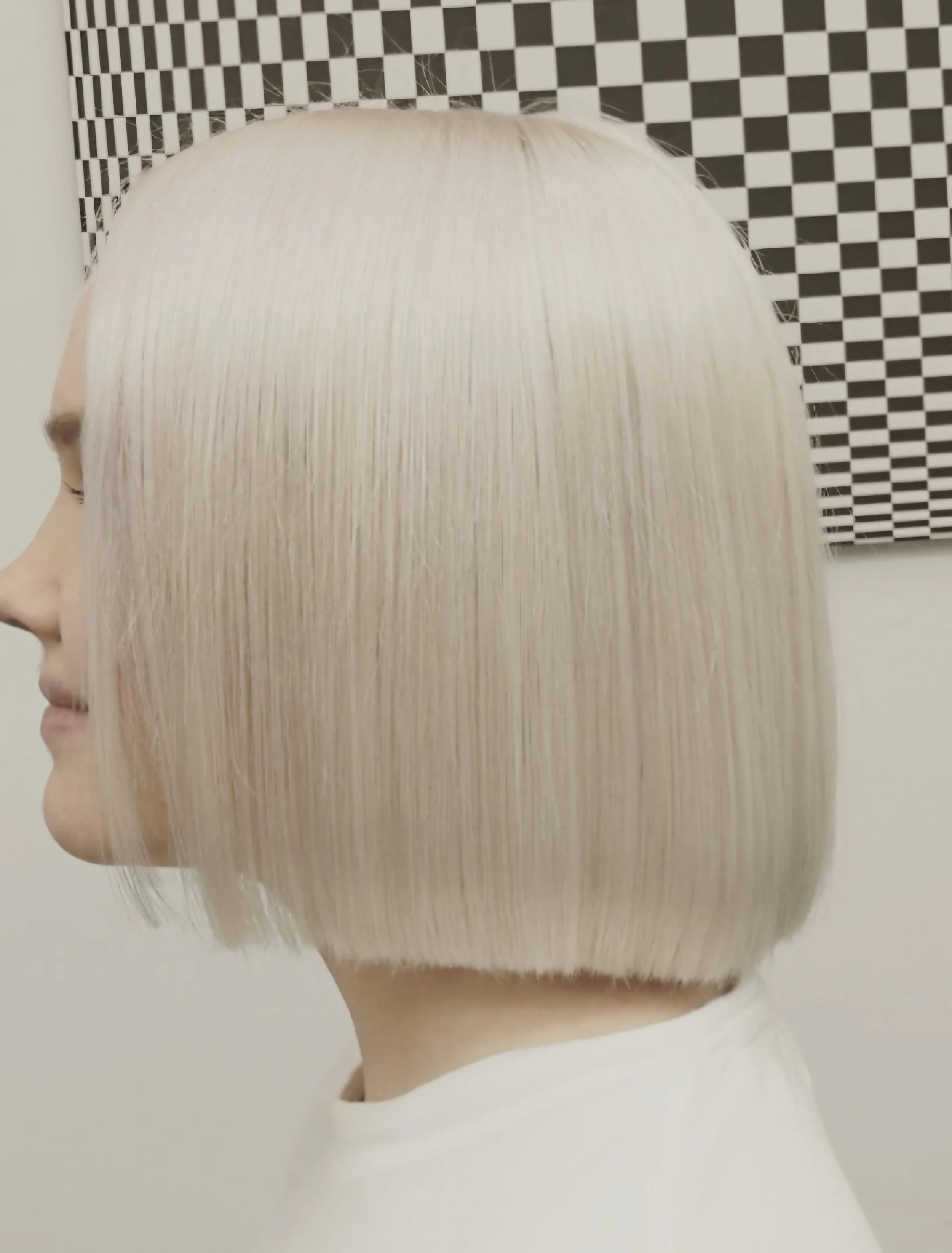 A mannequin with a short, platinum blonde bob hairstyle. The background features a black and white checkered pattern.