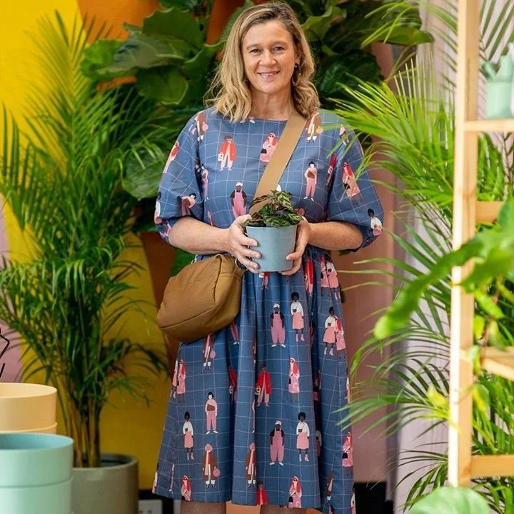 A person in a blue dress with a colorful pattern holding a potted plant, standing among various green plants with a yellow wall in the background.