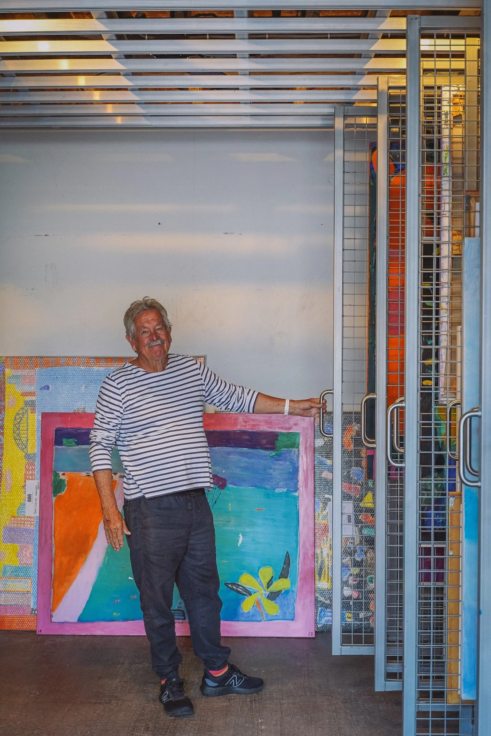 Man standing in an art storage room, leaning against colorful paintings on racks.