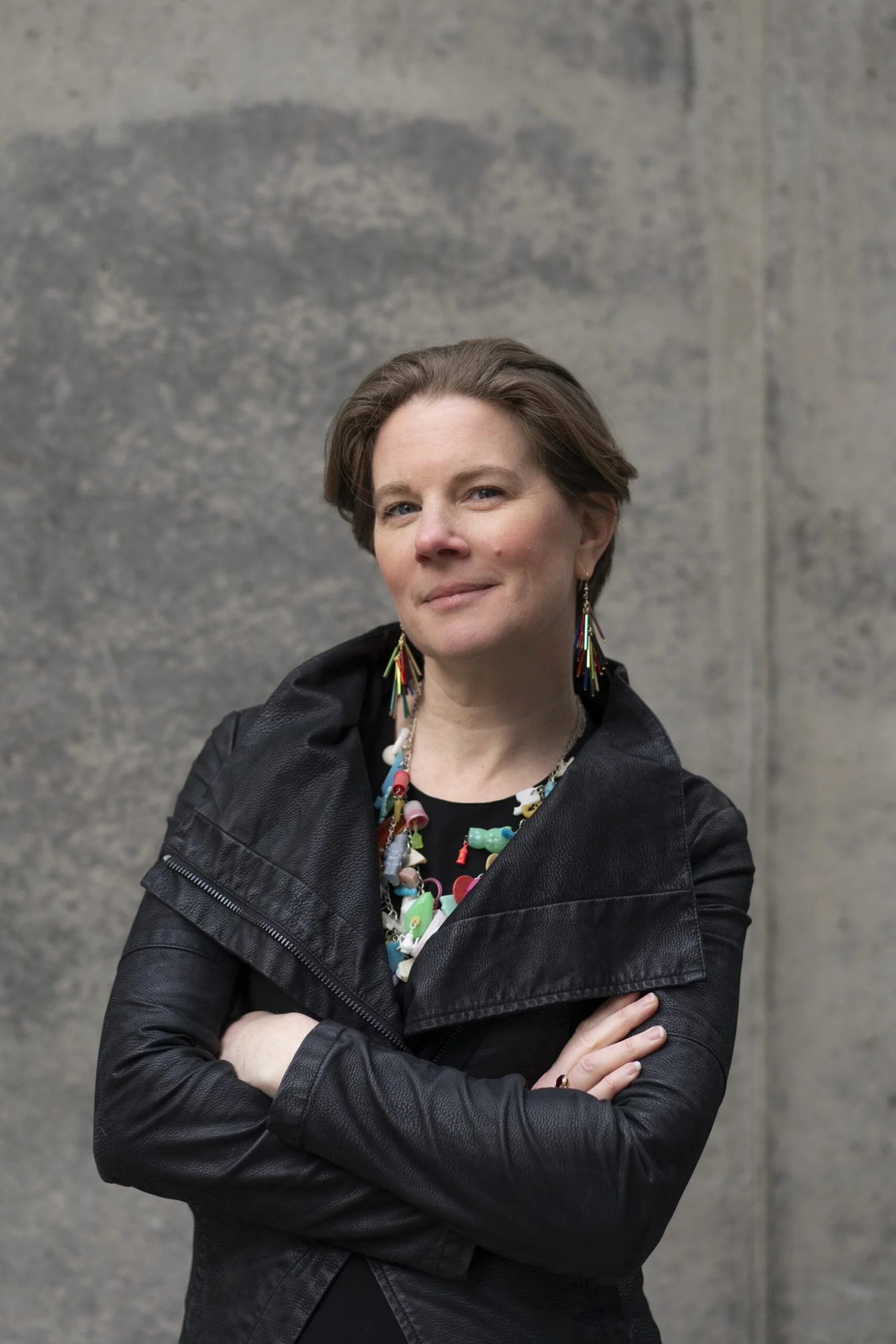 Person with short hair wearing a black leather jacket and colorful jewelry, standing with arms crossed in front of a gray concrete wall.