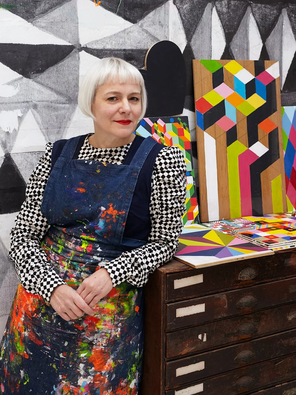 Artist in colorful paint-splattered apron standing in front of geometric abstract artworks and a monochrome wall mural.