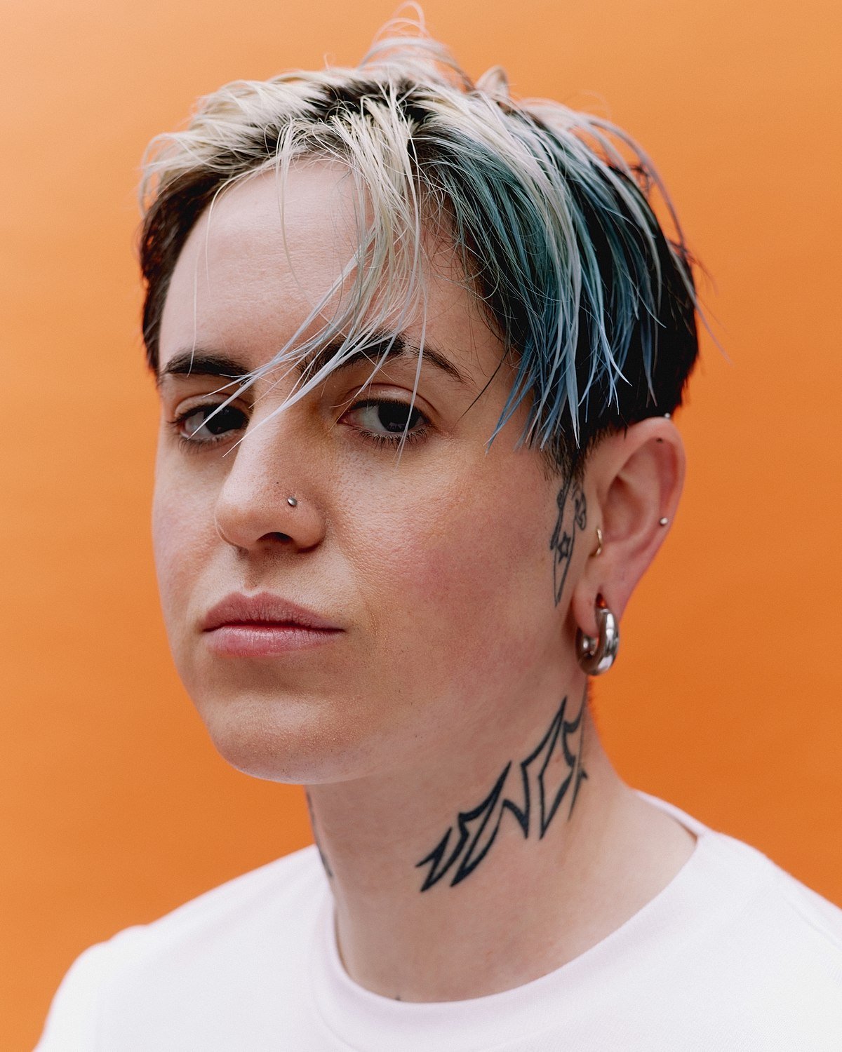 Person with short, two-toned hair, nose and ear piercings, and neck tattoo against orange background.