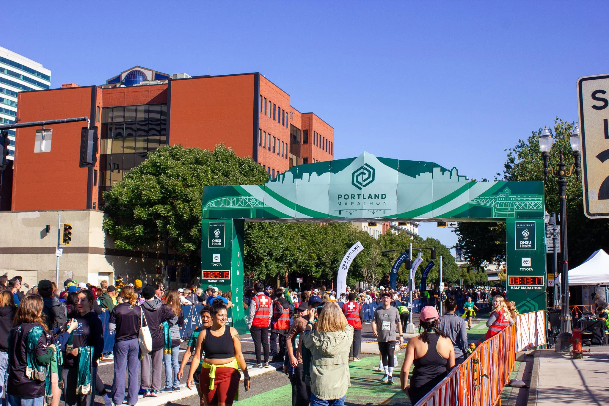 People gather at the Portland Marathon start line, with a large green Portland Marathon banner overhead, in an urban setting on a sunny day.