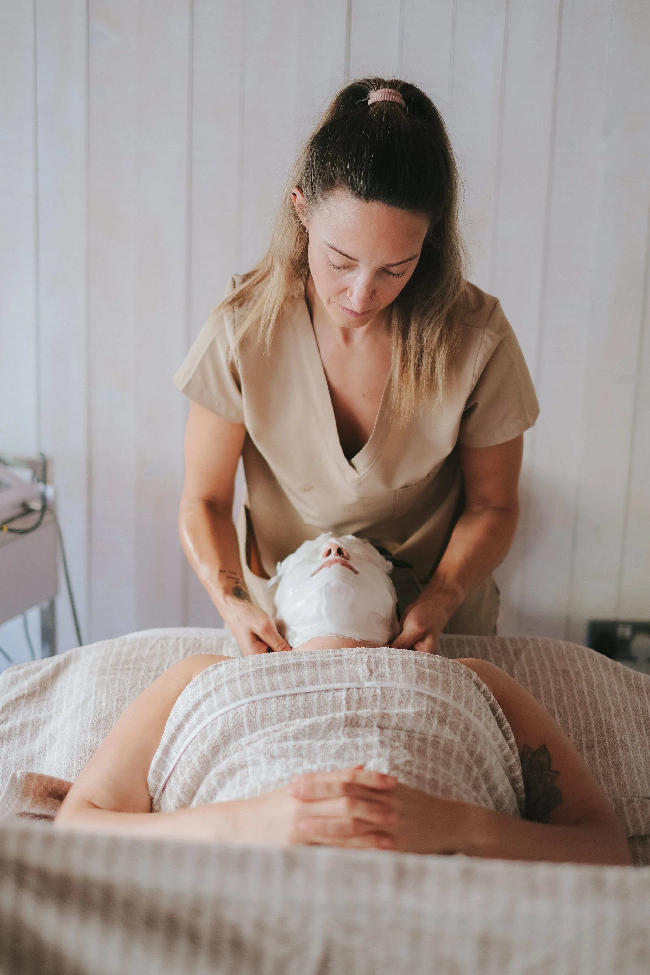 A woman lying on a massage table with a towel, receiving a facial treatment from a skincare specialist in a spa room.