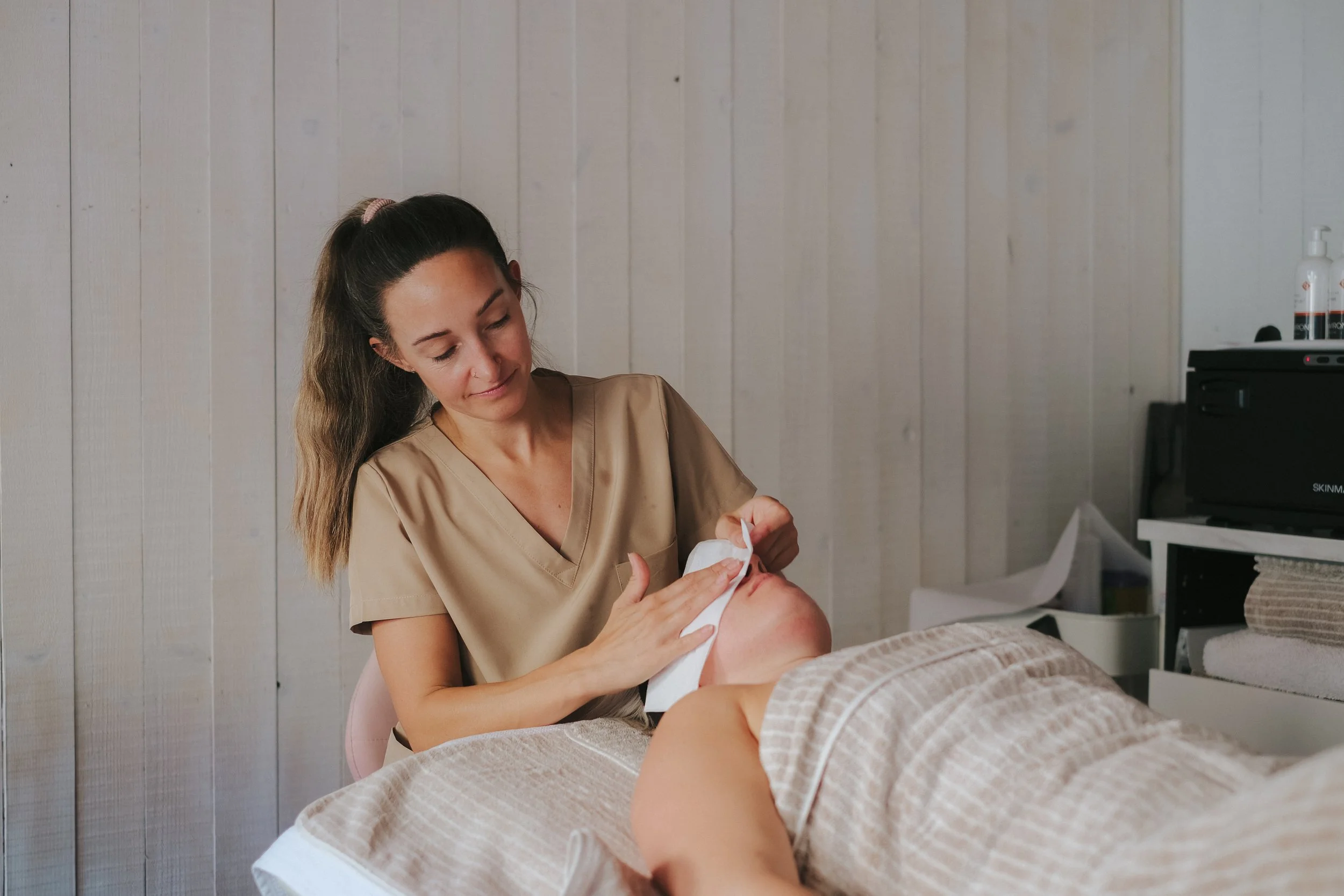 A female skin health specialist in a beige uniform tending to a client lying face-up on a treatment bed in a skincare clinic, with white panelled walls in the background.