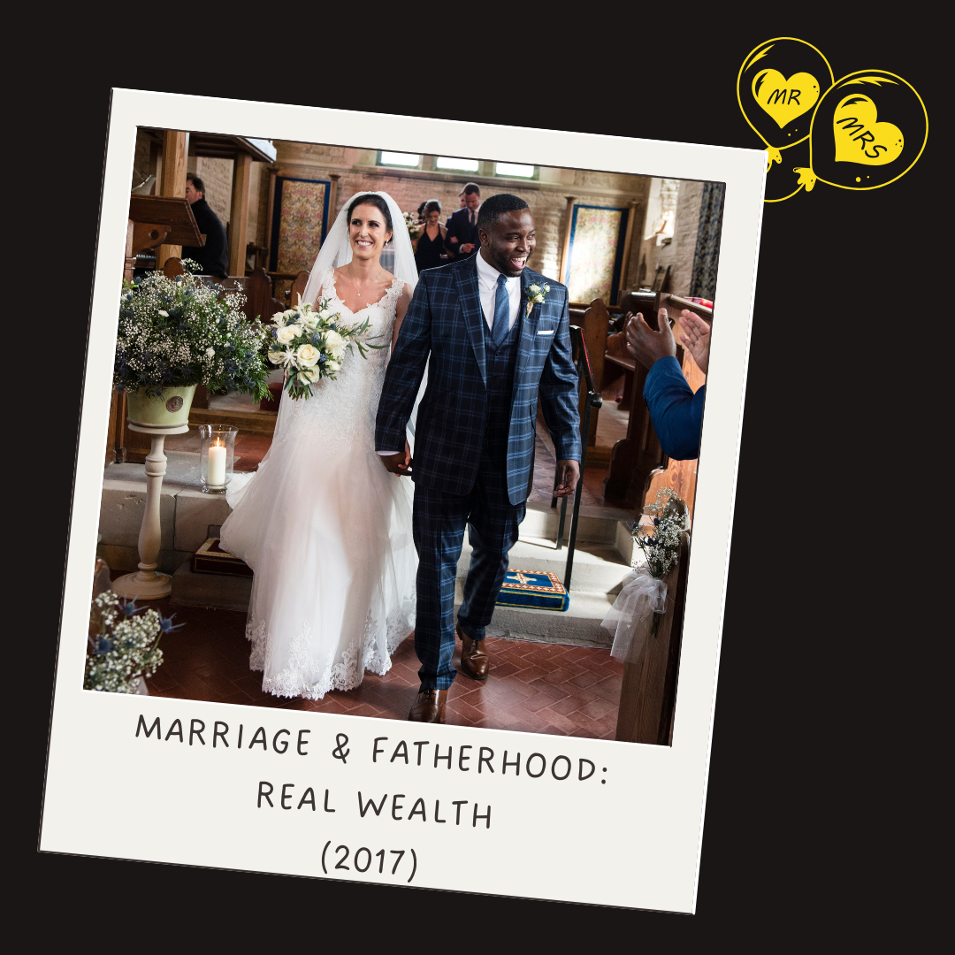 A bride and groom walk down the aisle after their wedding ceremony, holding hands and smiling. The bride wears a white lace wedding dress, and the groom wears a dark blue plaid suit. There are flowers and candles around them, and people are clapping in the background. The photo has a caption reading "Marriage & Fatherhood: Real Wealth 2017" with yellow balloons labeled "Mr" and "Mrs".