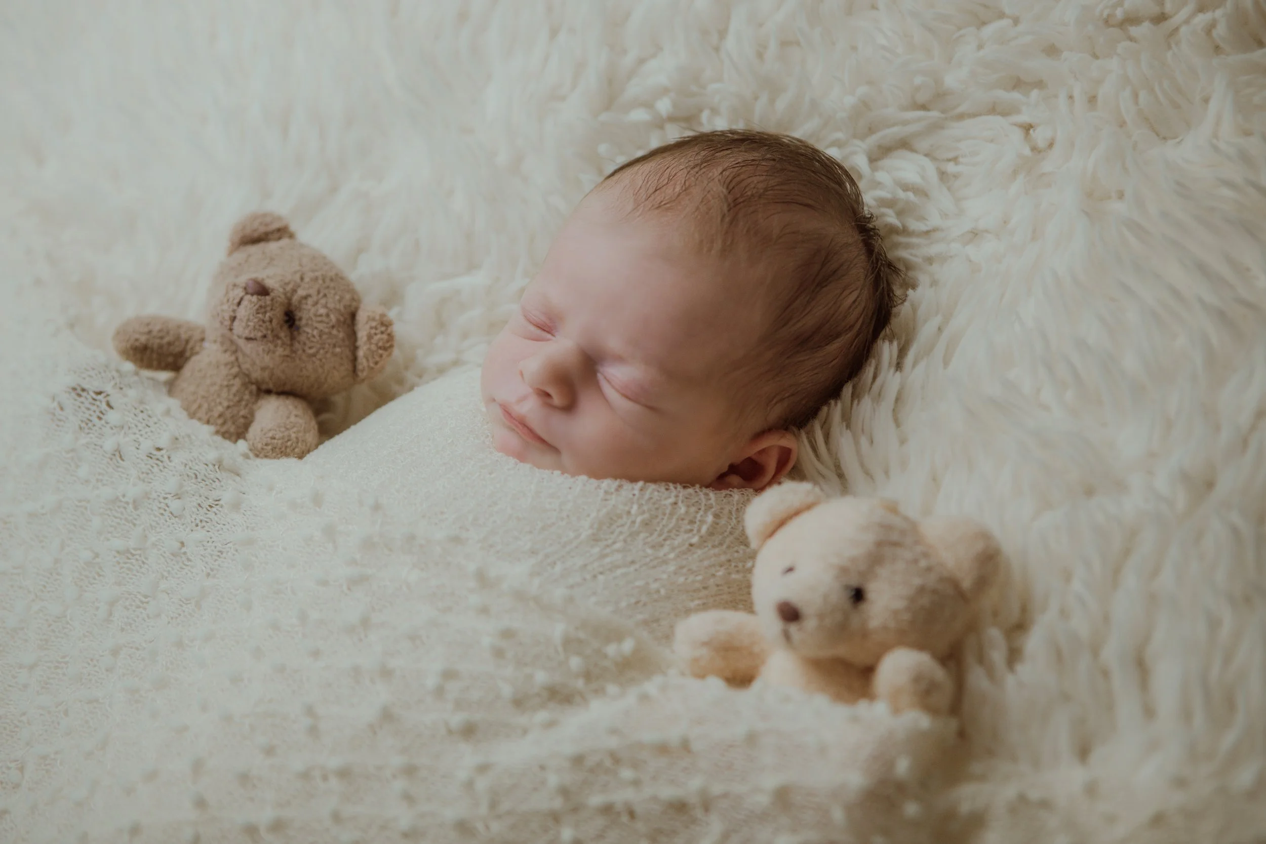 A sleeping baby lying on a soft, fluffy cream-colored surface, surrounded by two small beige teddy bears.