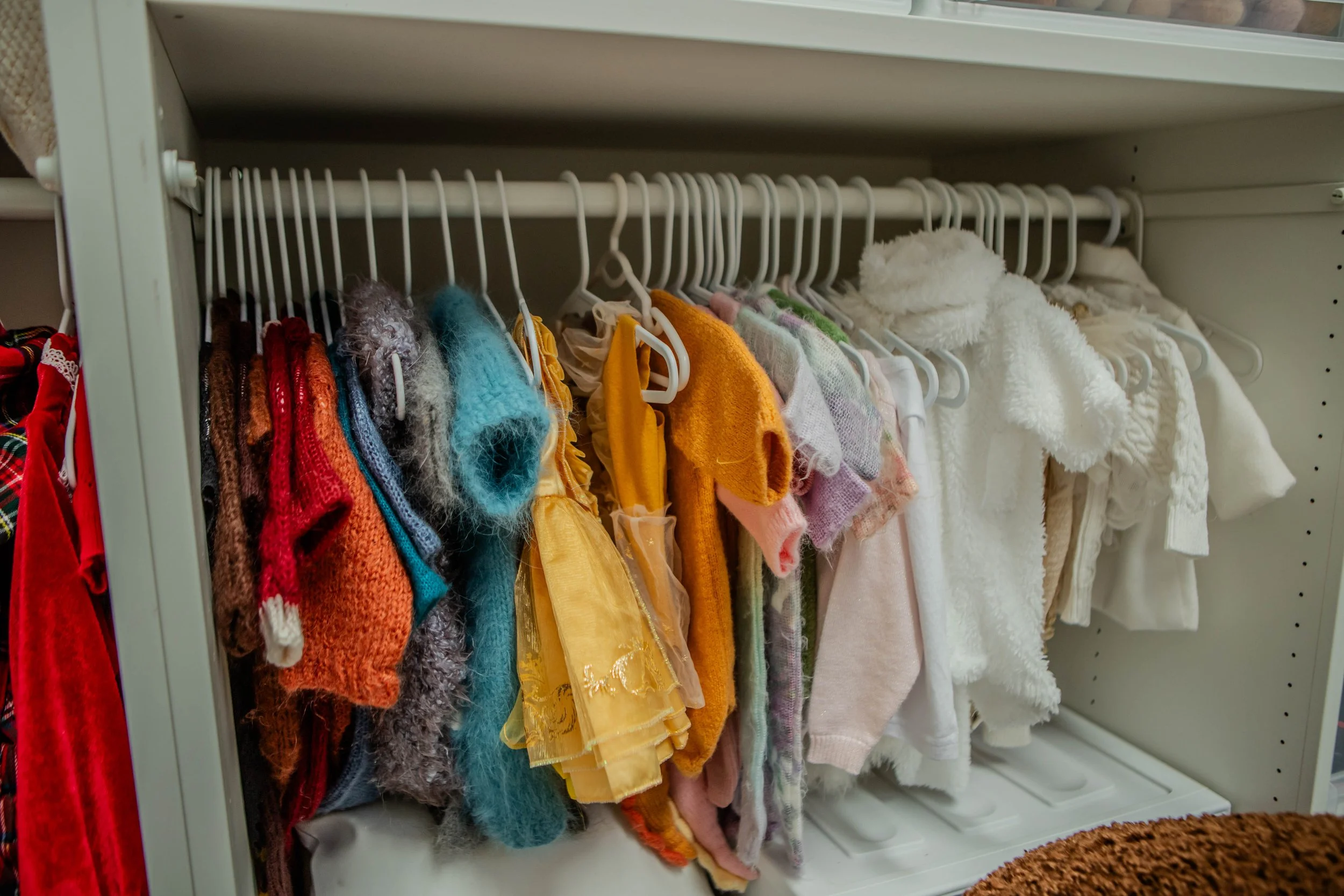 A closet shelf holding a variety of small, colorful, and fluffy doll clothes hanging on white hangers.