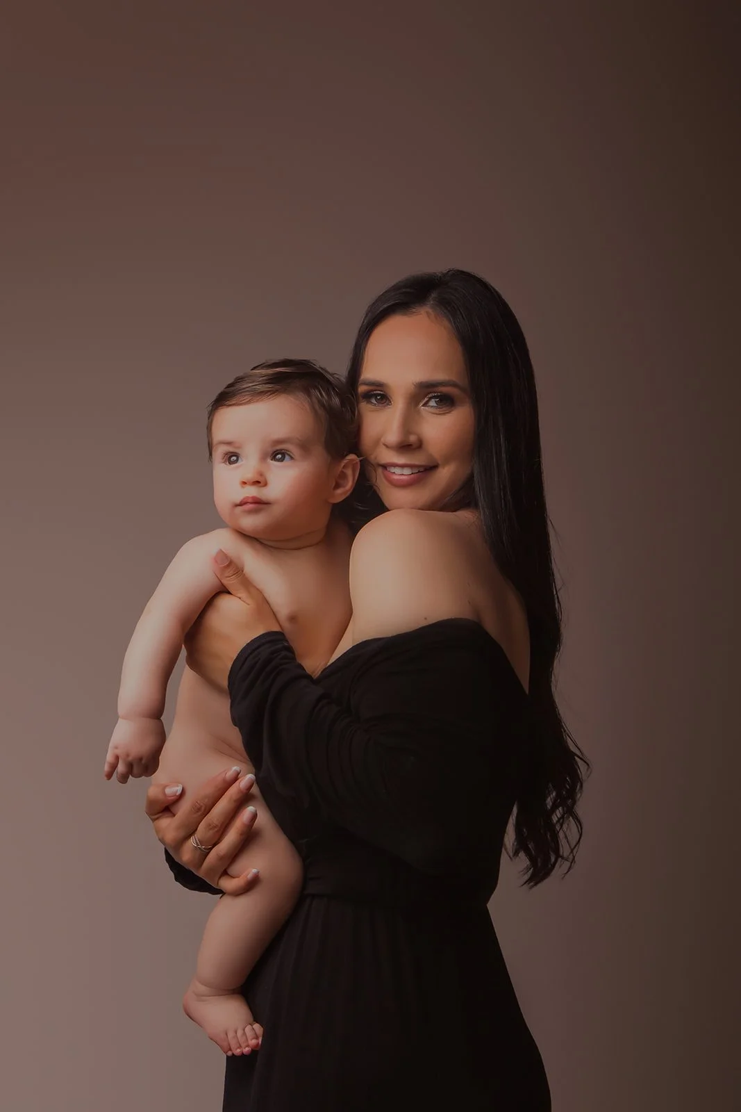A woman with long dark hair holding a smiling baby with blue eyes and light skin against a plain brown background.
