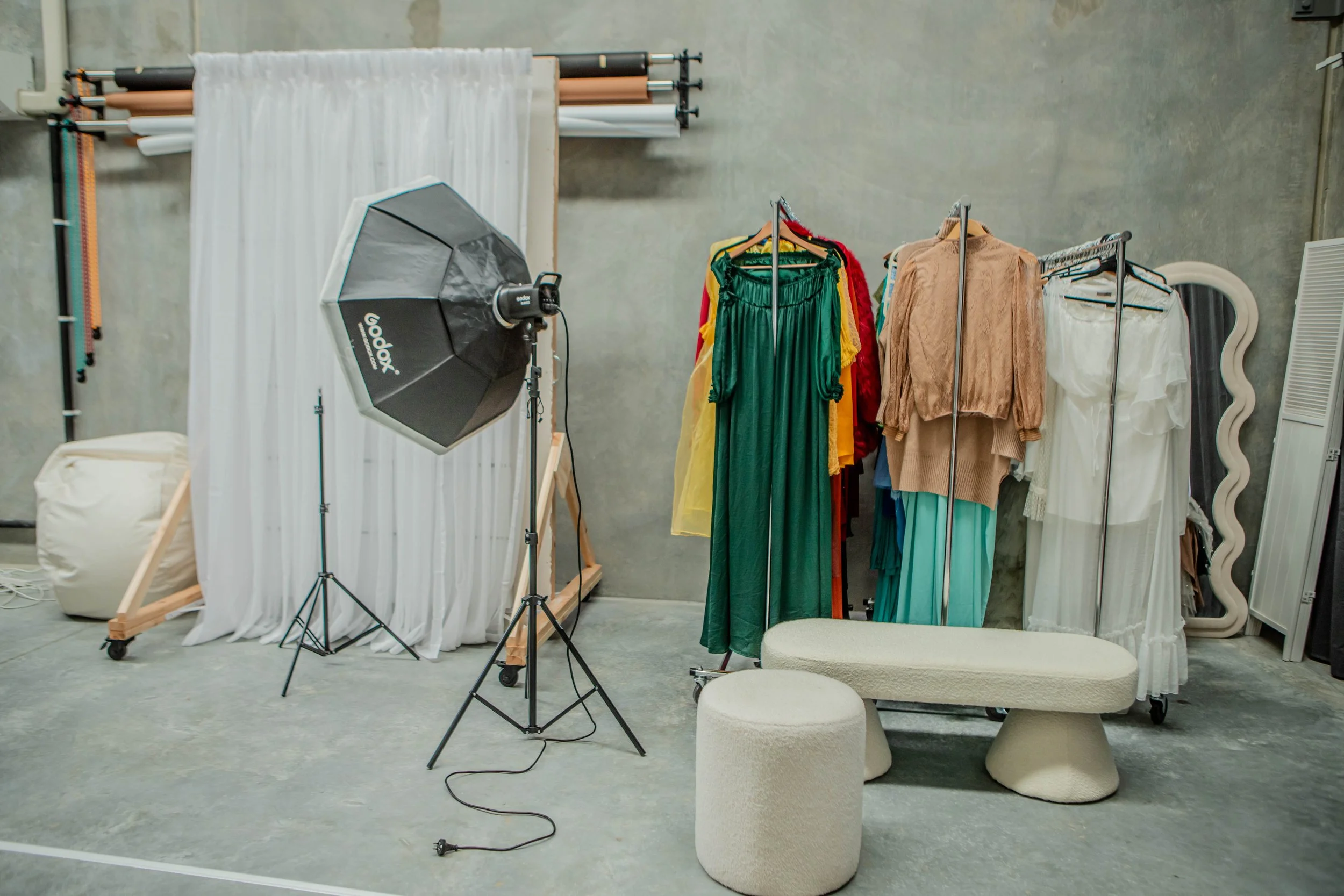 Photo of a photography studio setup with clothing racks, colorful dresses, a lighting umbrella, and a white backdrop.