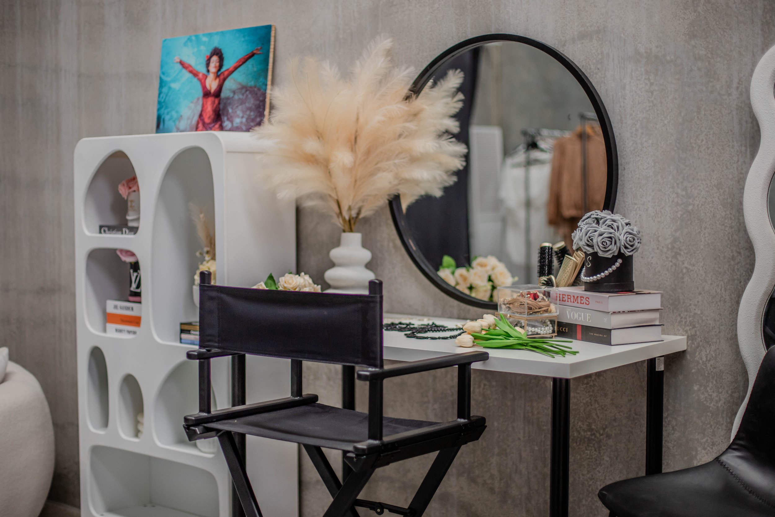 A vanity area with a mirror, white shelf, and black chair; decorated with a bouquet of gray roses, books, and a vase of pampas grass.