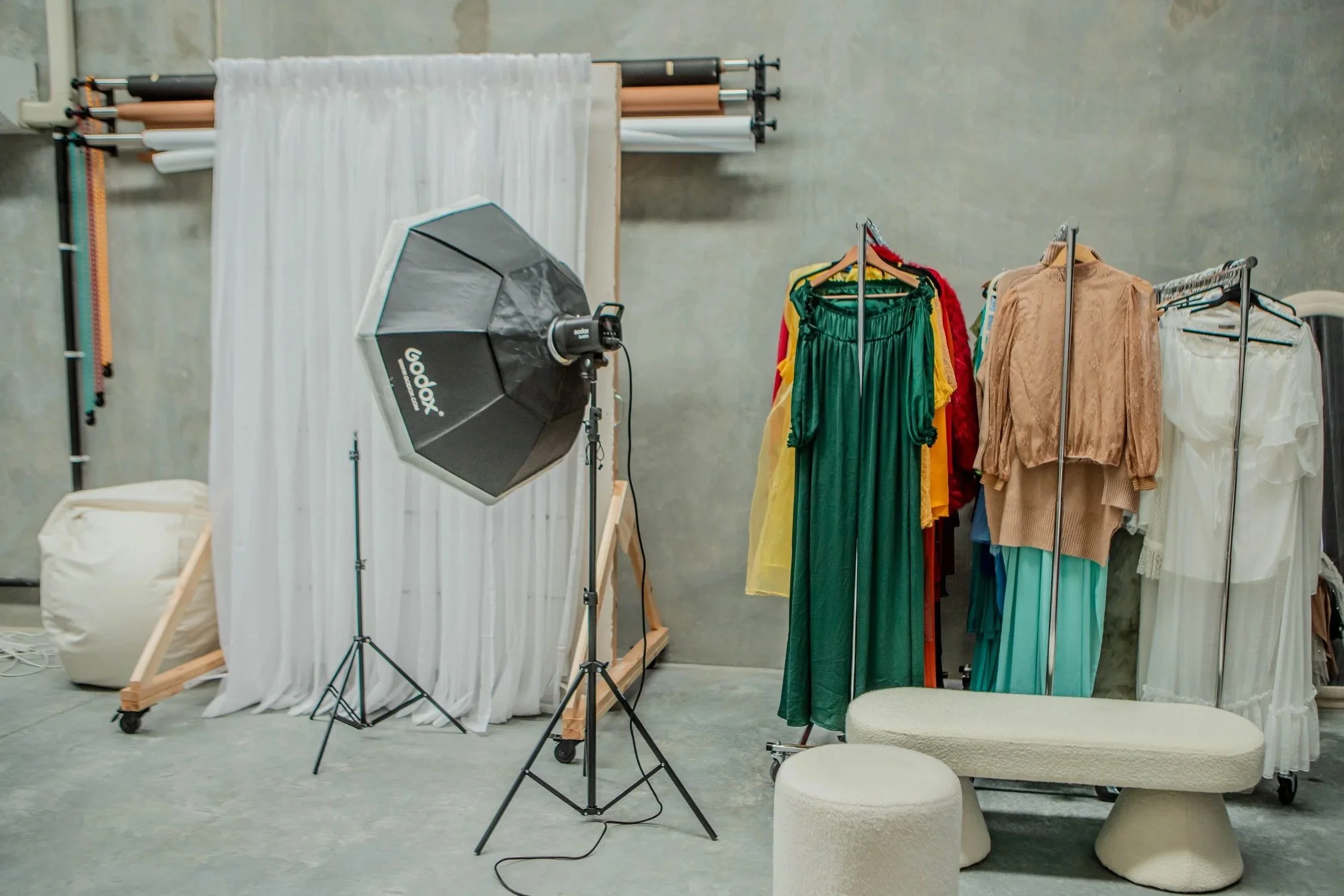 Studio setup with clothing racks featuring dresses and tops, a white fabric backdrop, professional lighting equipment, and seating furniture in a concrete-walled space.
