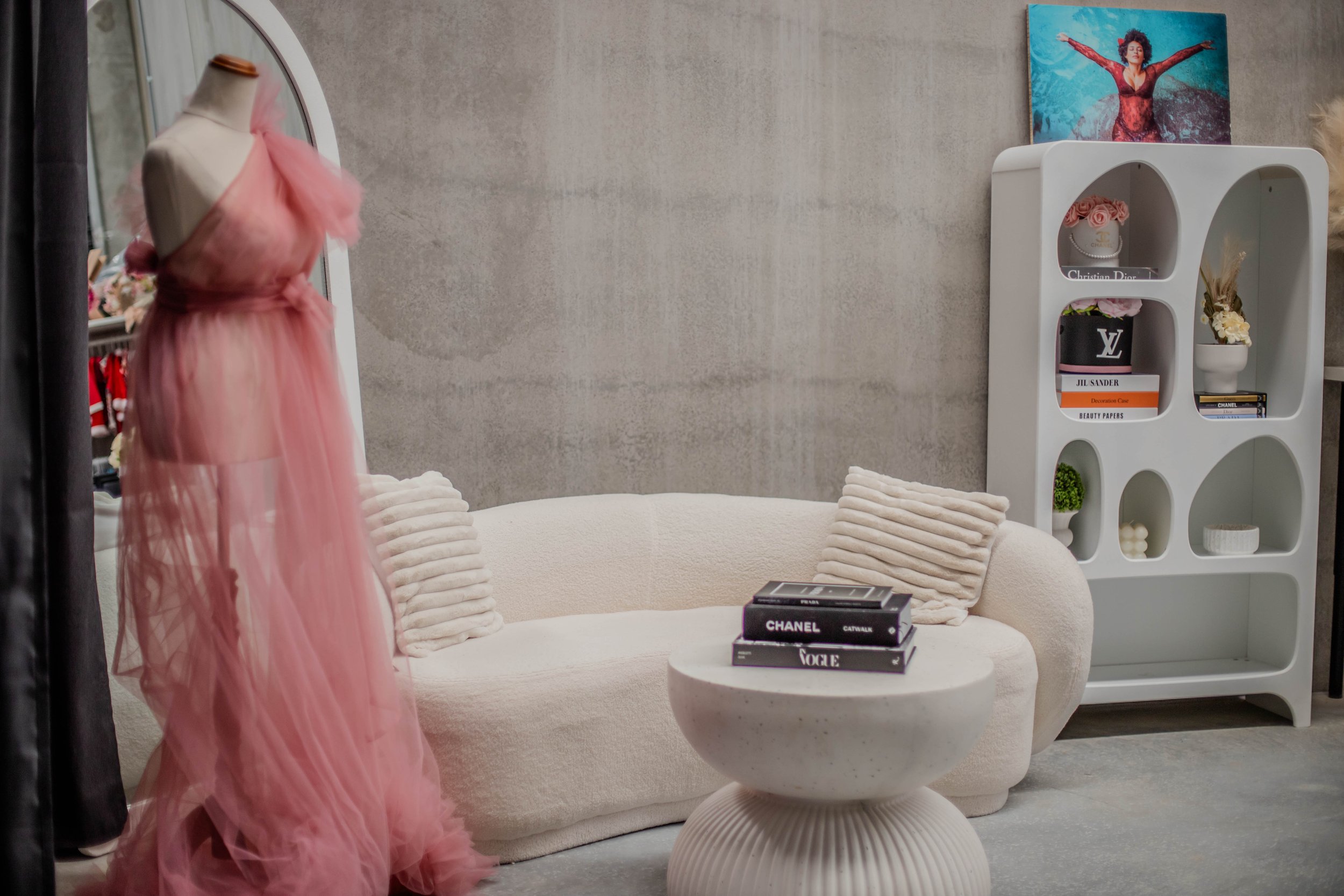A fashion boutique with a pink tulle dress on a mannequin, a cream-colored sofa with striped pillows, a round white coffee table with fashion magazines, and a white shelving unit displaying designer items and books. A colorful painting is on the wall behind the shelving unit.