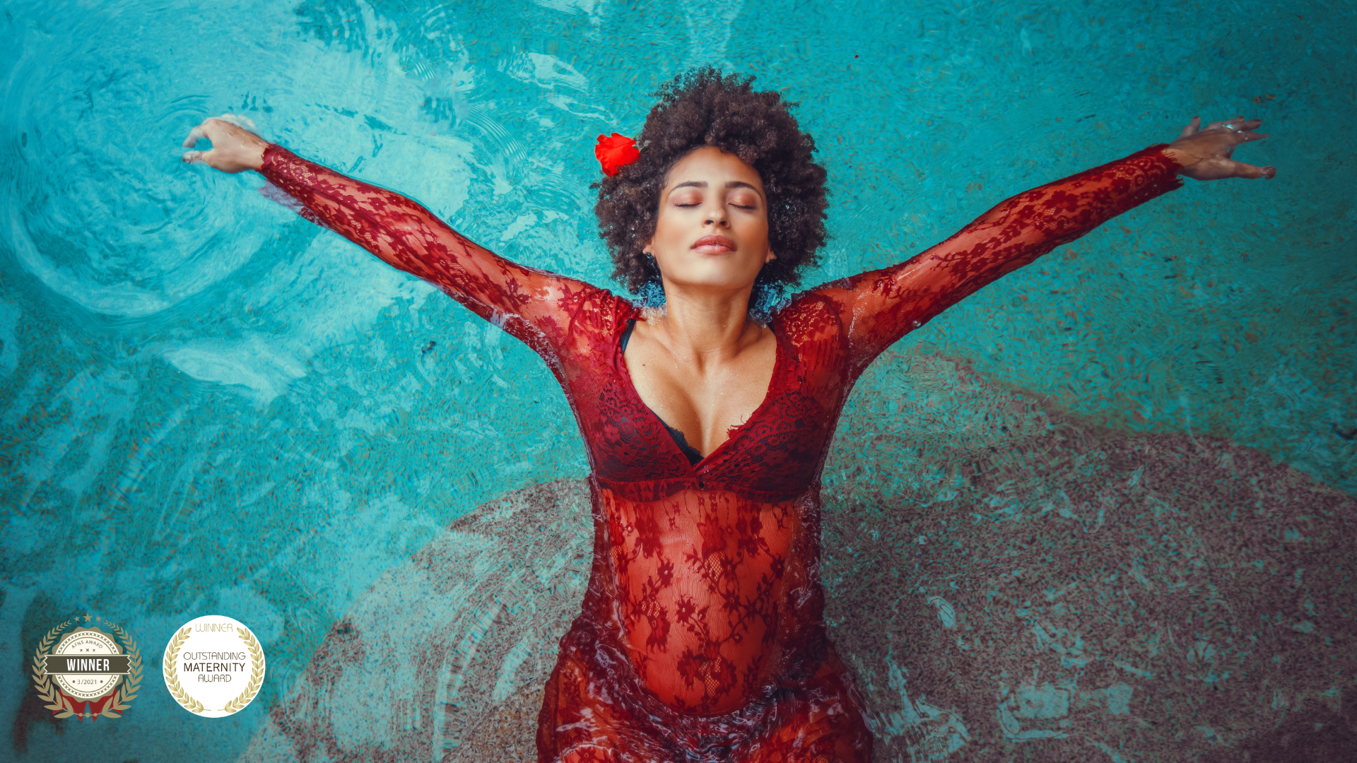 Woman in a red lace dress floating with arms outstretched in a turquoise pool, eyes closed, wearing a red flower in her curly hair.