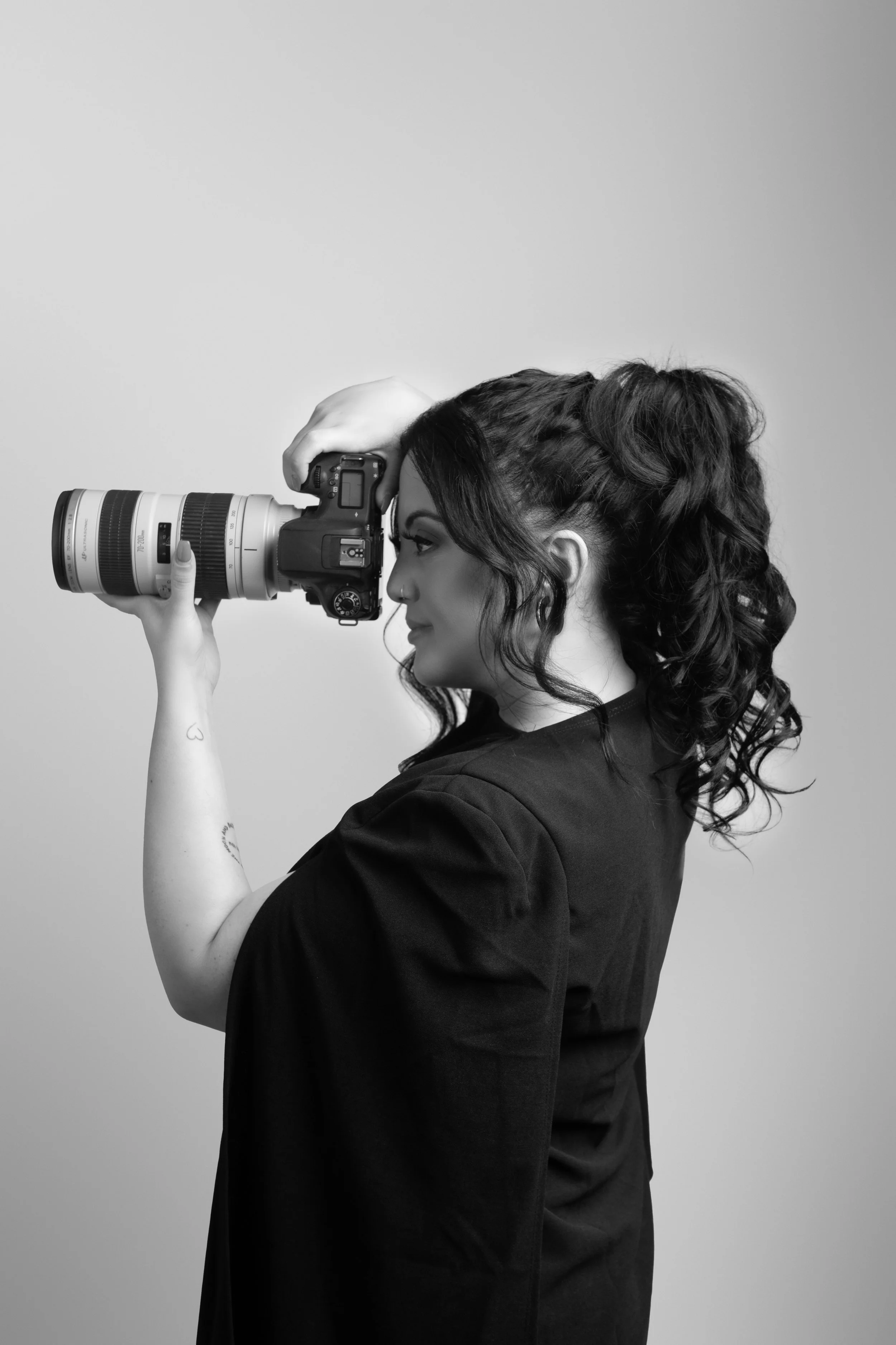 A woman with curly hair in a black shirt holds a professional camera with a long lens up to her face, looking through the viewfinder.
brisbane award winning photographer Studio Sabrina Luz Rothwell