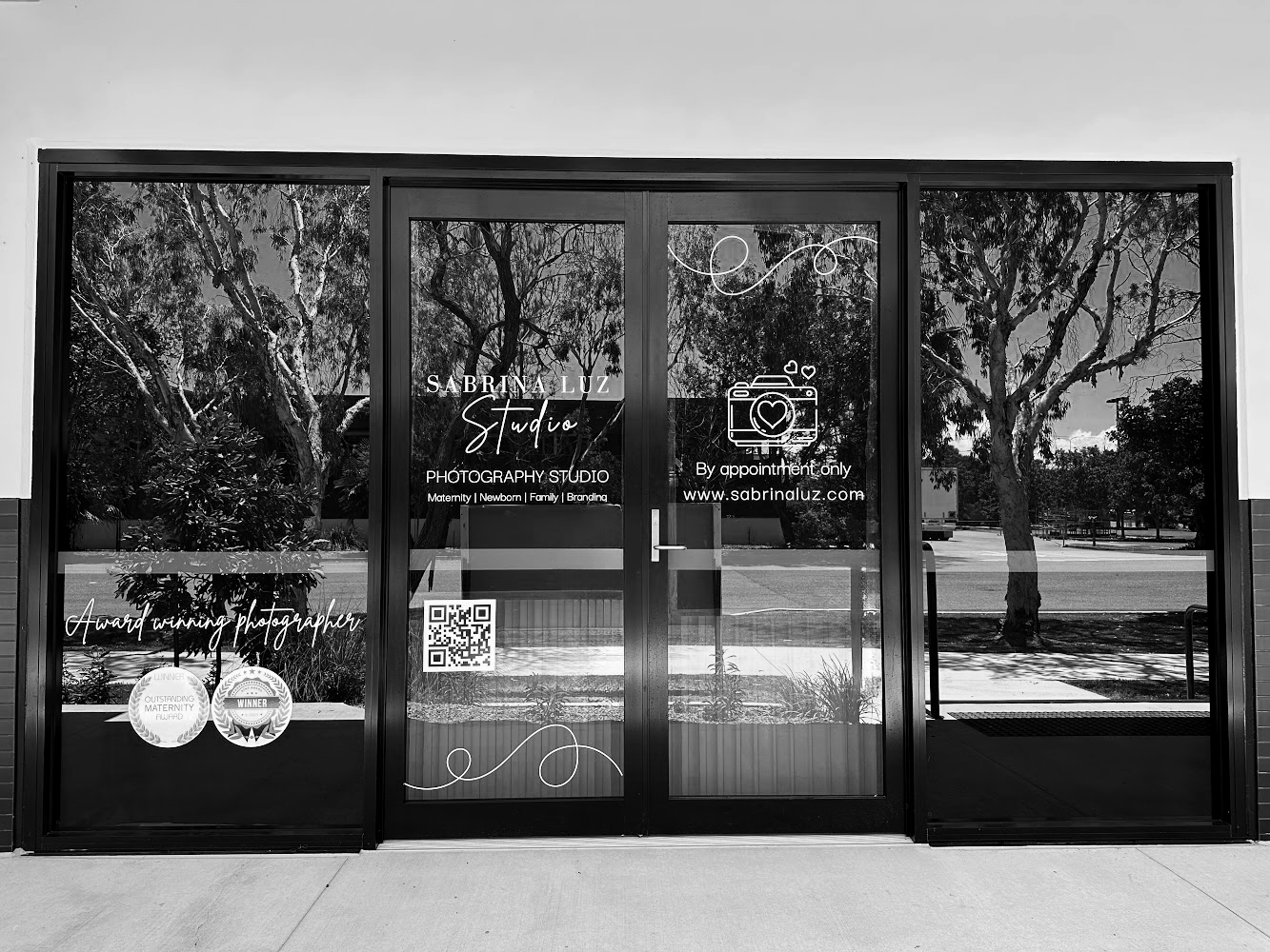 Glass door with photography studio branding, including text about Sabrina Luz Studio, awards, and services, with a QR code and outdoor reflections of trees and sky.