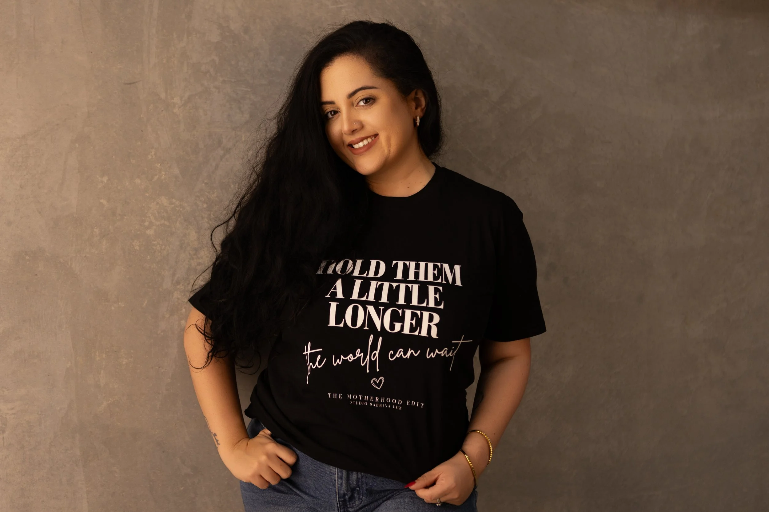 A woman with long dark hair wearing a black T-shirt with white text and dark jeans, standing against a textured gray wall. She is smiling and has her left hand on her hip.