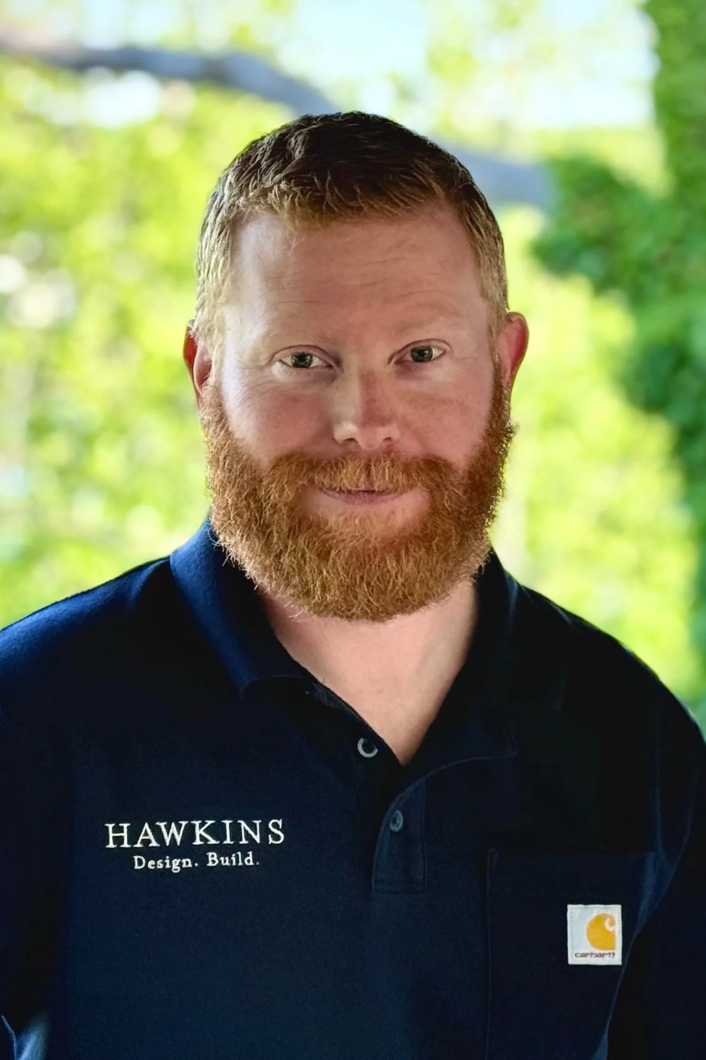 Portrait of a man with red hair and a beard wearing a dark carhartt shirt with brands Hawkins and Carhartt, standing outdoors with green trees in the background.