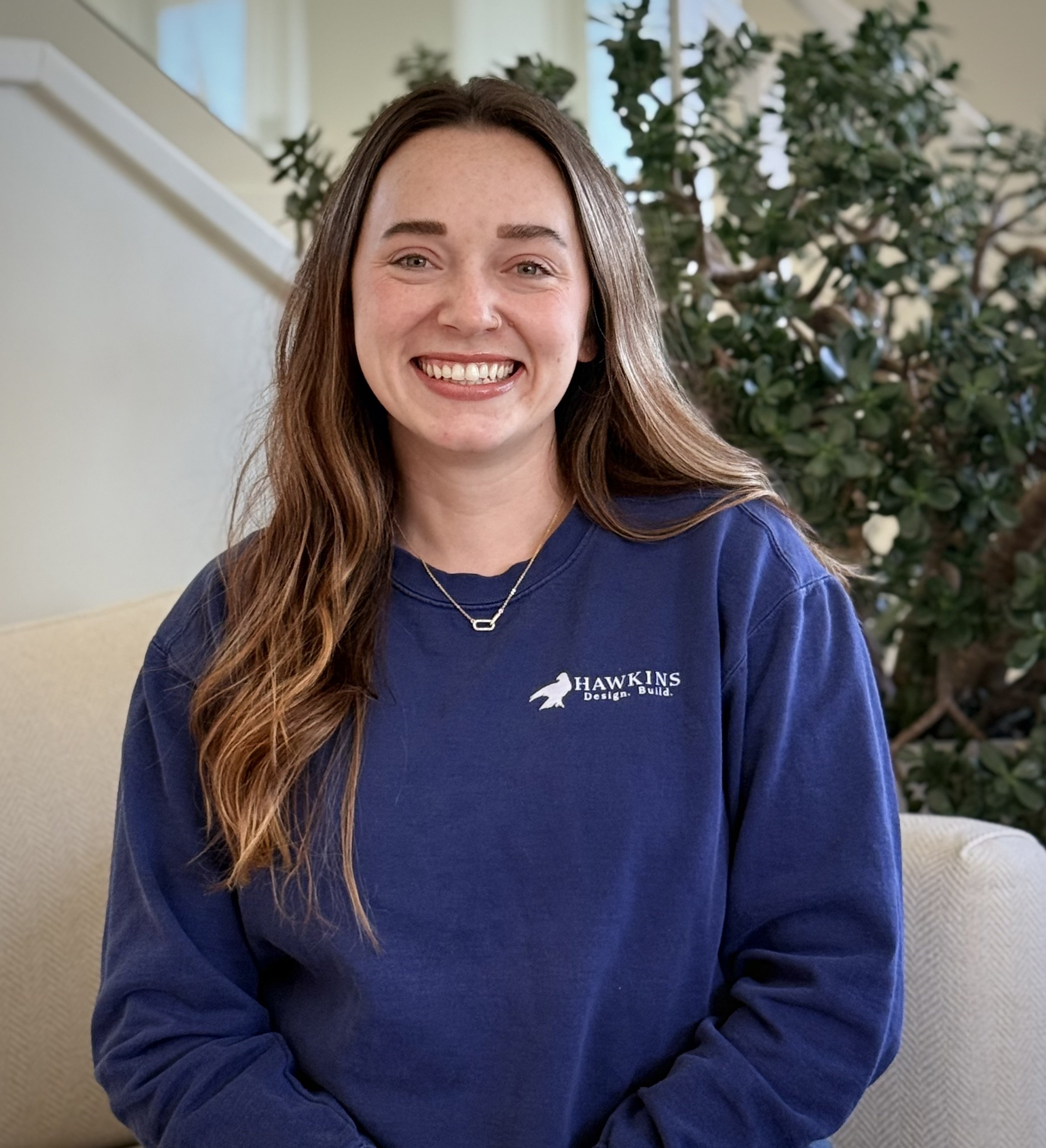 A young woman with long dark hair, smiling, wearing a black top with ruffled sleeves and a necklace, against a dark background.