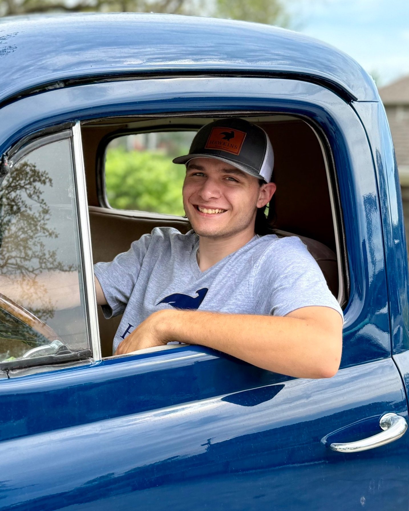 Smiling young man with a cap sitting in the driver's seat of a blue vintage truck, with trees and a house in the background.