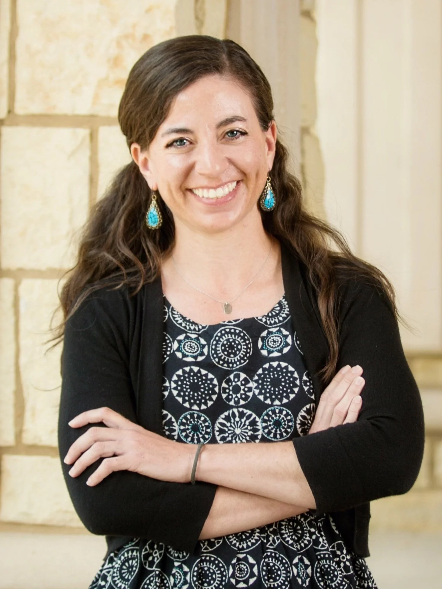 A smiling woman with long wavy brown hair, wearing a black and white patterned dress, turquoise earrings, and a necklace, standing with her arms crossed against a beige brick wall.