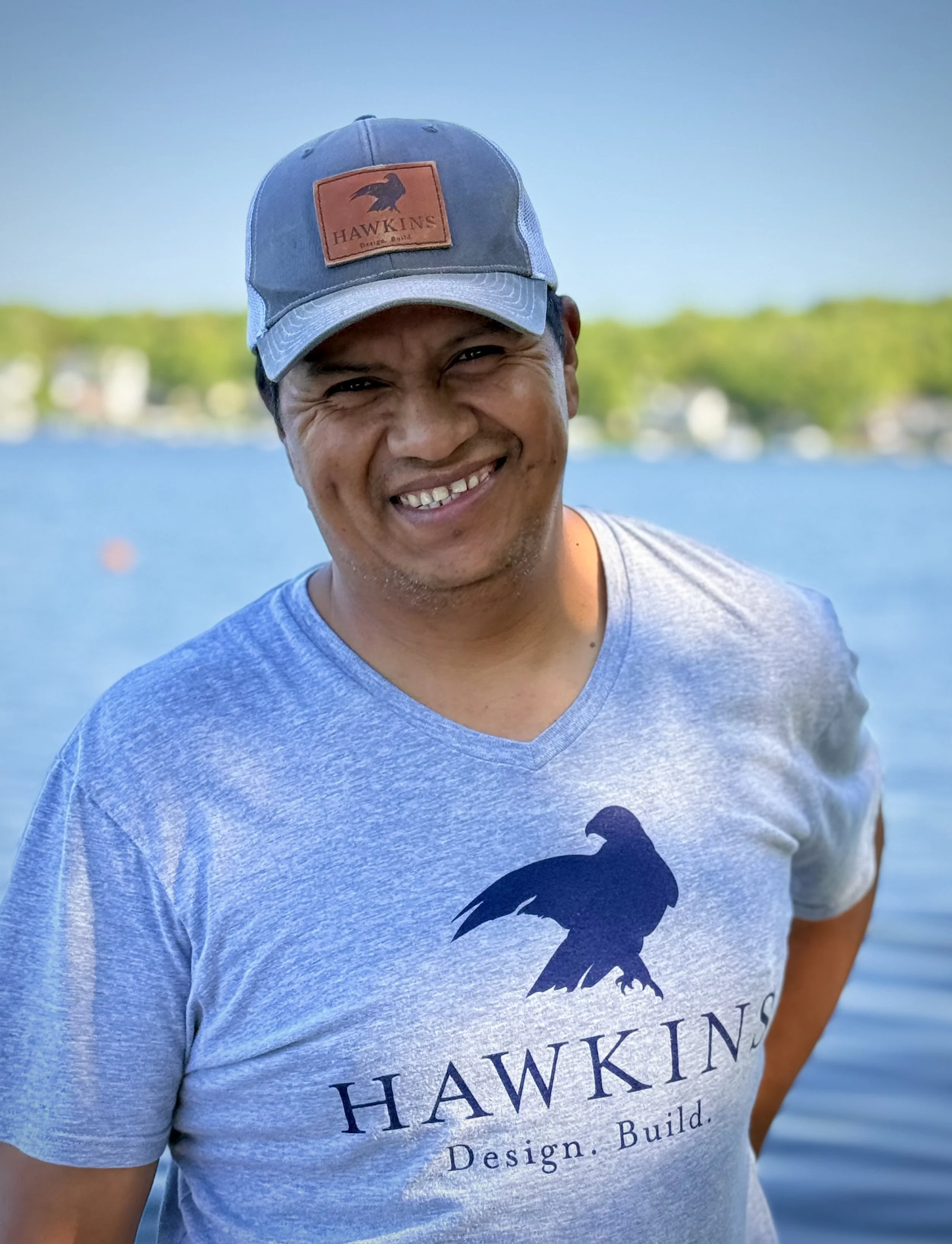 Smiling man wearing a blue Hawkins cap and a gray T-shirt with a black bird logo, standing outdoors near a body of water with trees in the background.