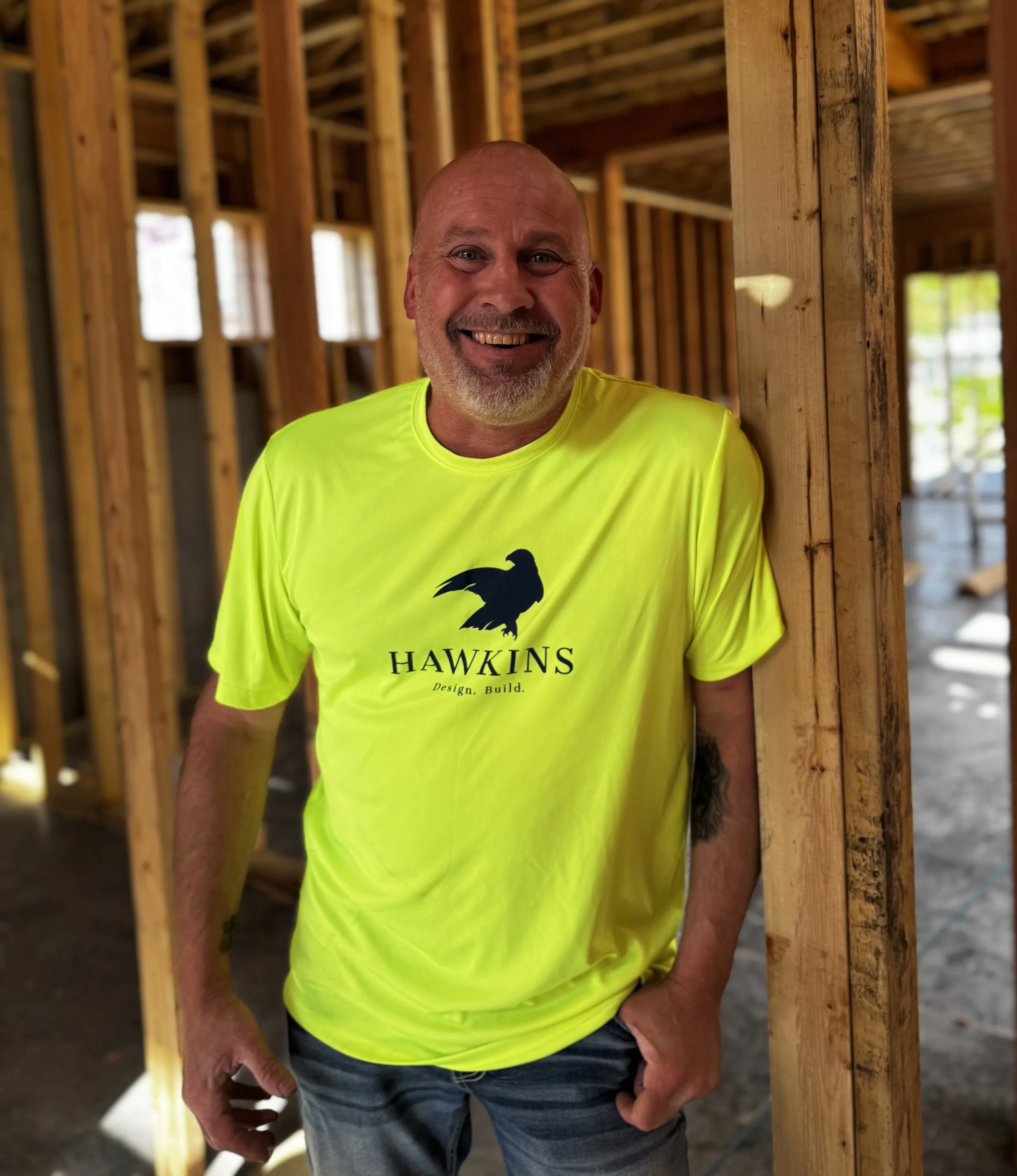 Man standing inside a wooden building under construction, wearing a bright yellow Hawkins t-shirt and smiling.