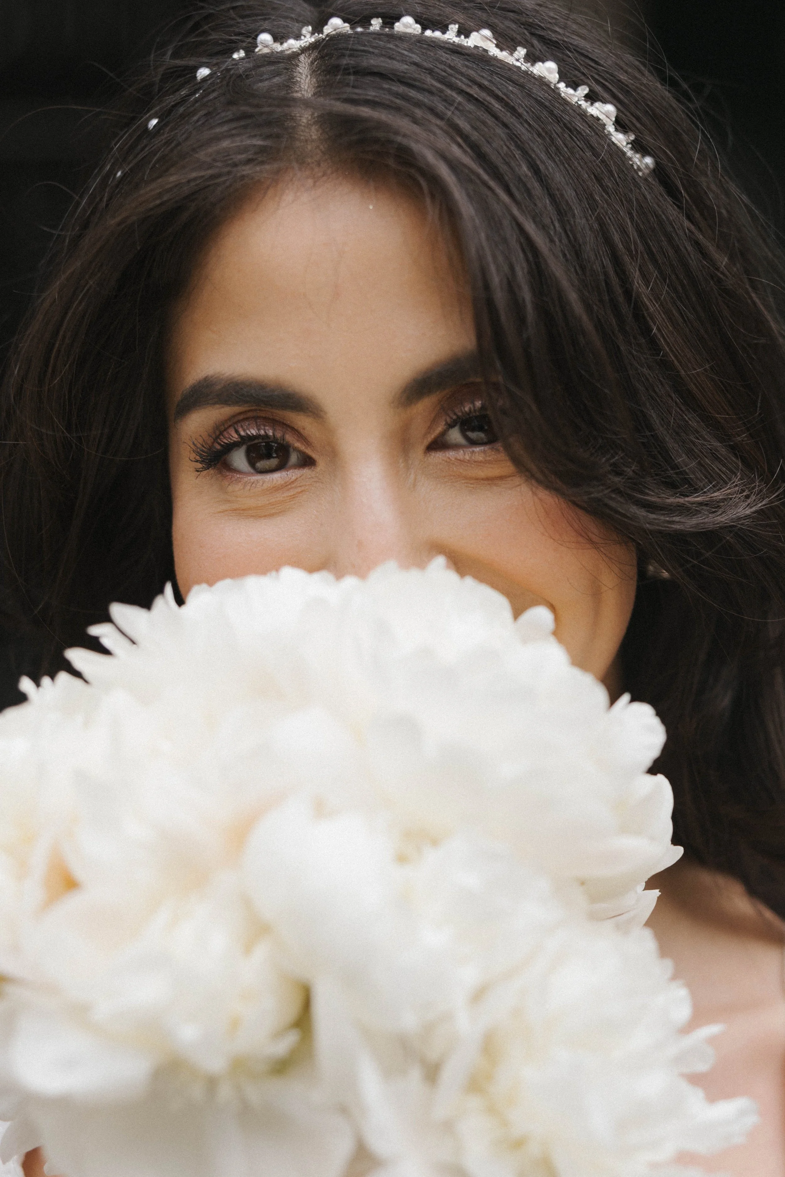 Nahaufnahme eines lachenden Mädchens mit dunklen Haaren, einem Diadem, das ihre Haare schmückt, und einem großen weißen Blumenstrauß, der ihr Gesicht teilweise verdeckt.