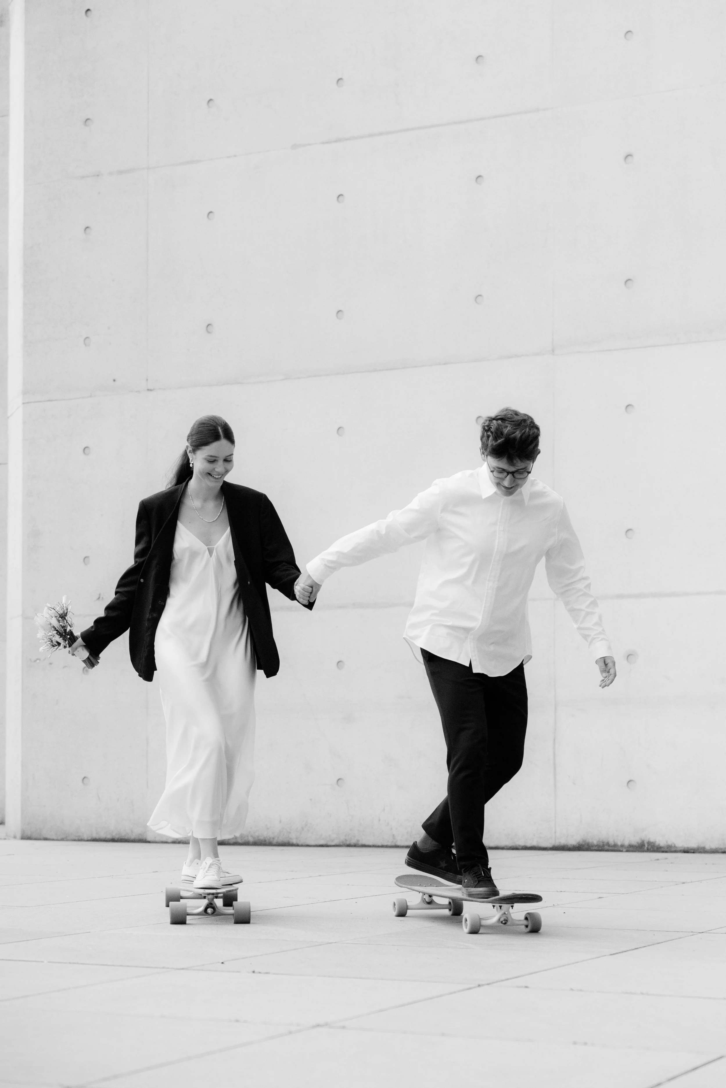 Ein junges Paar, das auf Rollern vor einer modernen Betonwand unterwegs ist, lächelt und hat Spaß.