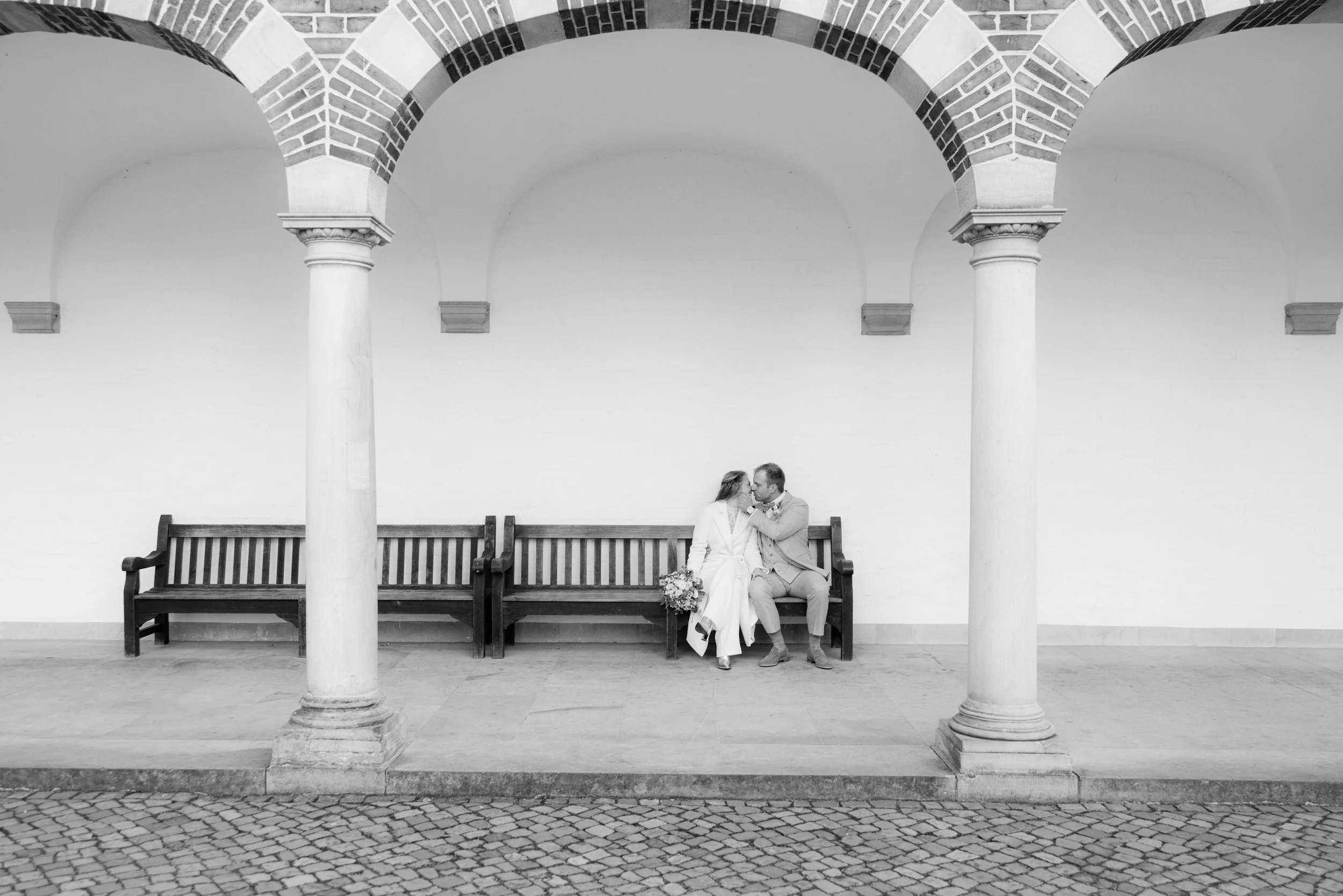 Ein Brautpaar sitzt auf einer Bank unter einer Arkade, küsst sich in schwarzer und weißer Fotografie.