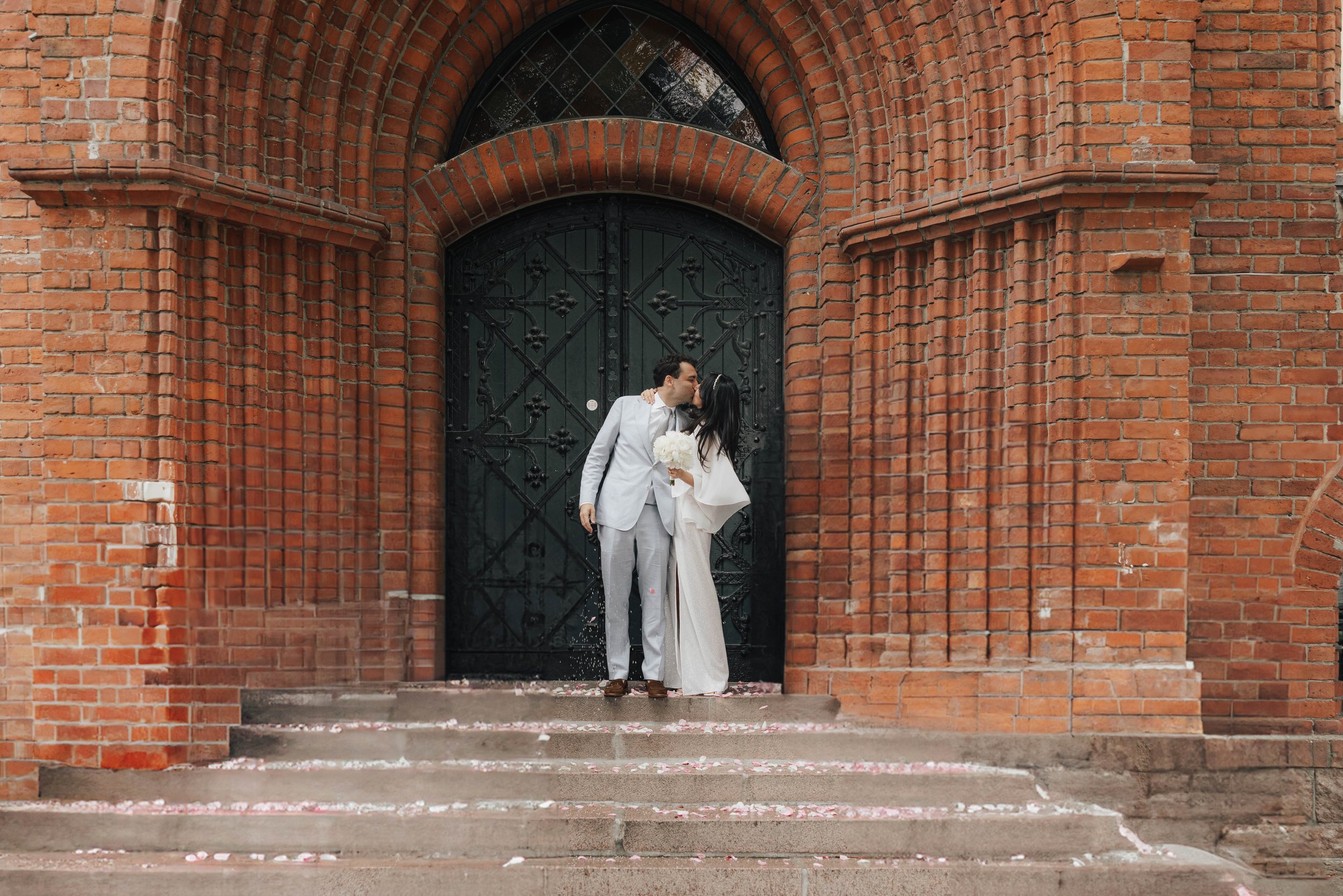Ein nach hochzeitskleidung gekleidetes Paar küsst sich vor einer großen schwarzen Tür an einer roten Backsteinmauer.
