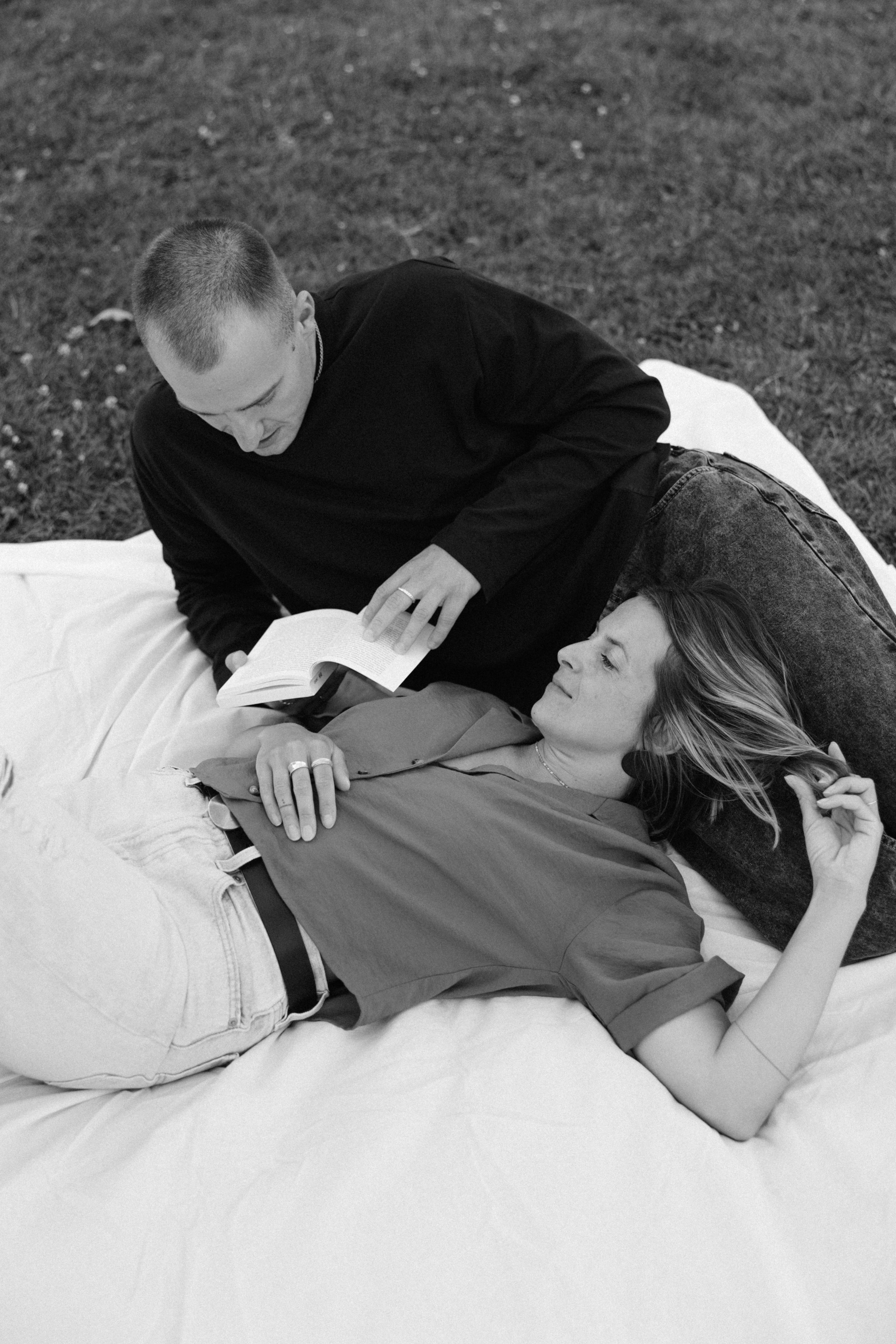 Ein Mann liest einer Frau ein Buch vor, während sie auf einer weißen Decke im Freien liegt.