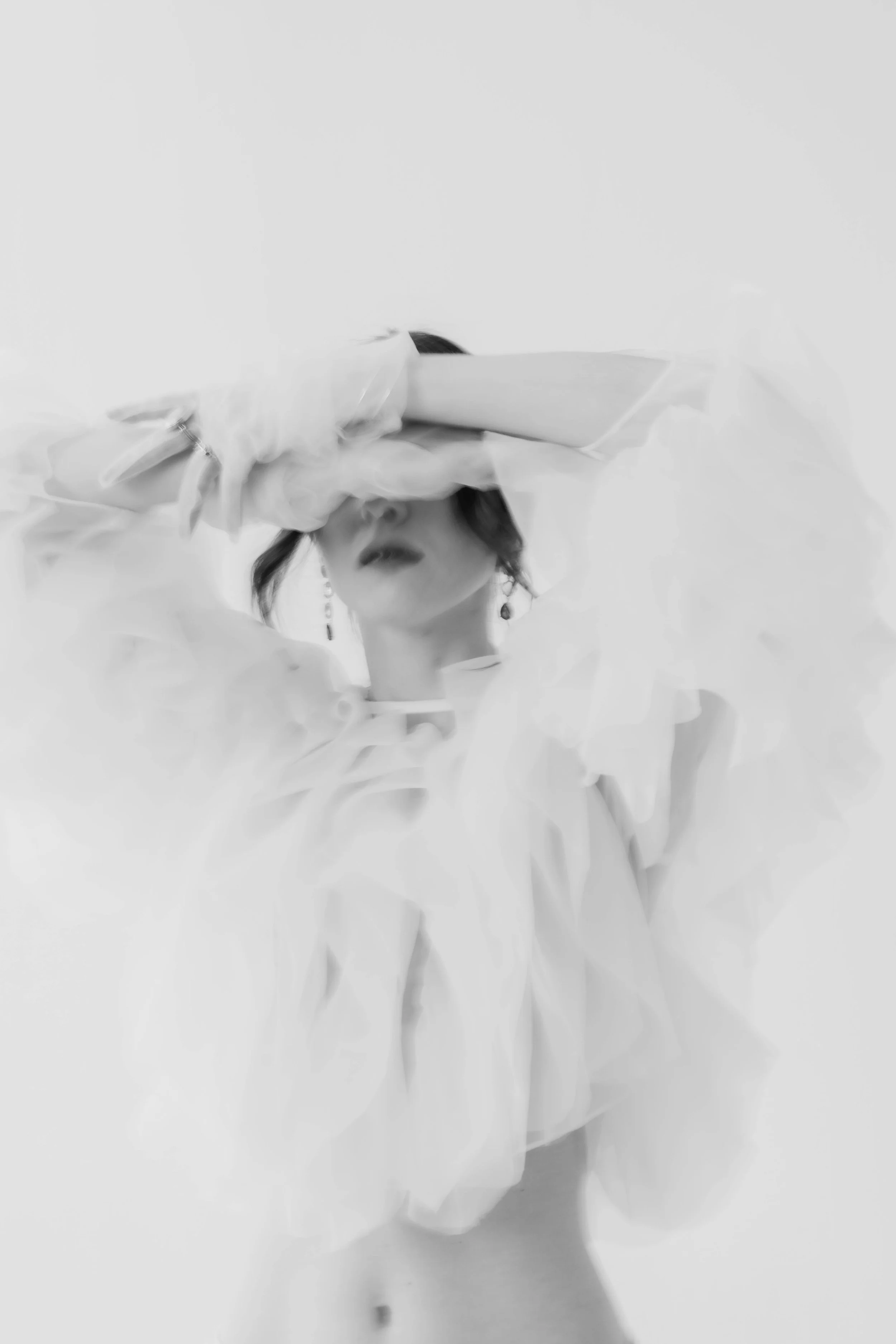 Schwarz-weiß-Foto einer Frau mit dunklen Haaren, die ein weißes, voluminöses Kleid trägt. Sie hält die Arme vor das Gesicht, trägt einen Handschuh und auffälligen Ohrring, der nur teilweise sichtbar ist.