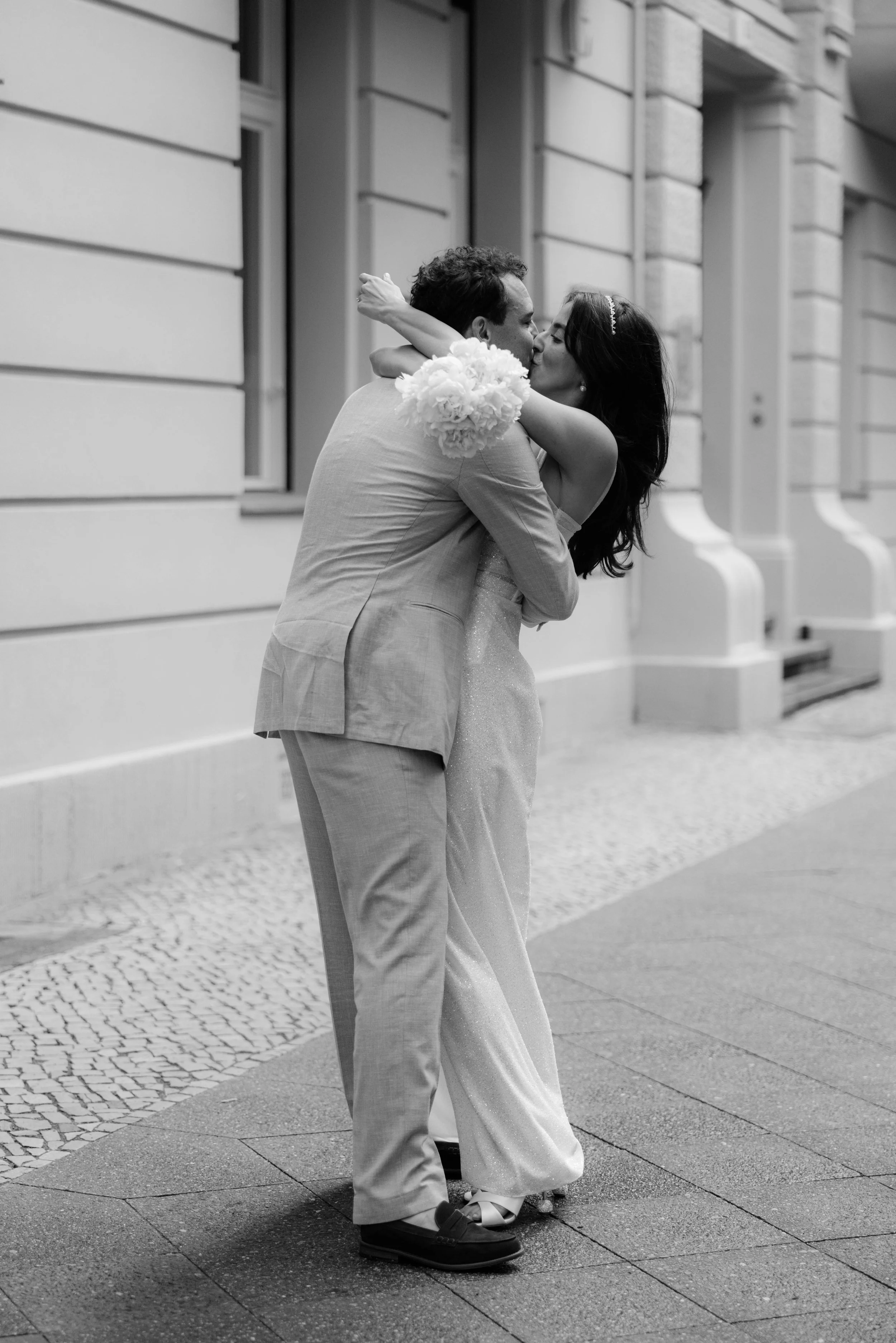 Ein Paar küsst sich leidenschaftlich auf der Straße, es scheint ein romantischer Moment bei einem besonderen Anlass zu sein, möglicherweise eine Hochzeit, in Schwarz-Weiß fotografiert.