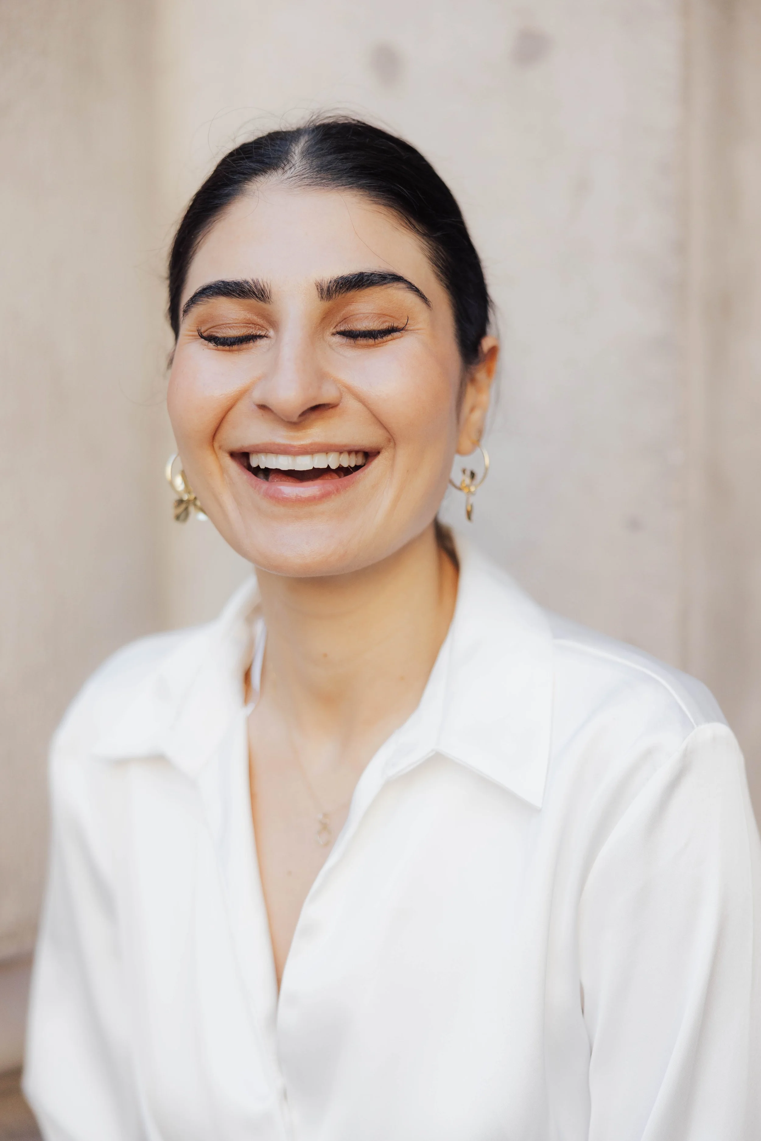 Junge Frau mit dunklem Haar, die lächelt, trägt goldene Ohrringe und ein weißes Hemd.