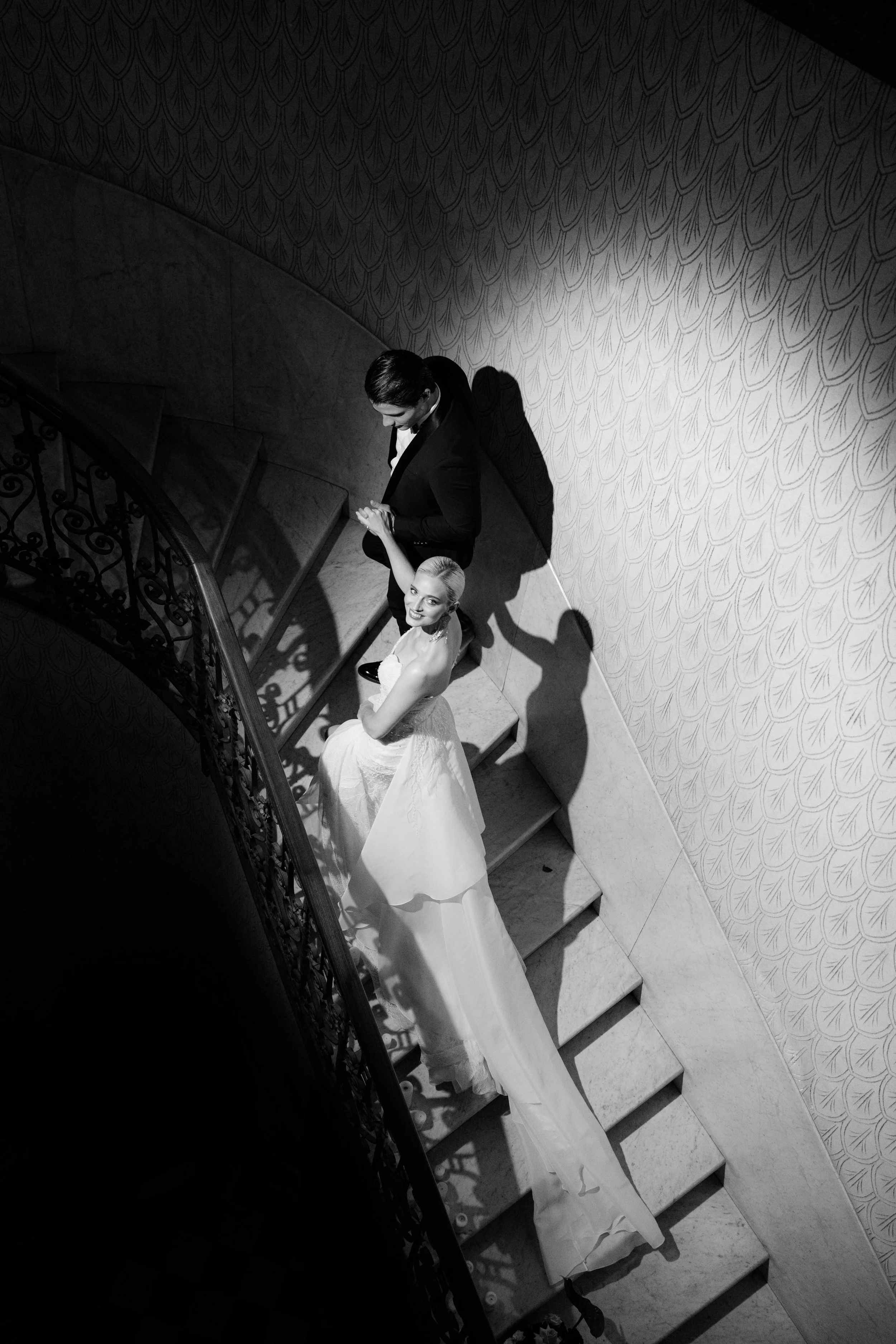 Schwarz-Weiß-Foto eines Paares in formeller Kleidung auf einer Wendeltreppe, die Dame trägt ein langes, voluminöses Kleid, der Herr einen Anzug. Sie halten Händchen und schauen sich an.