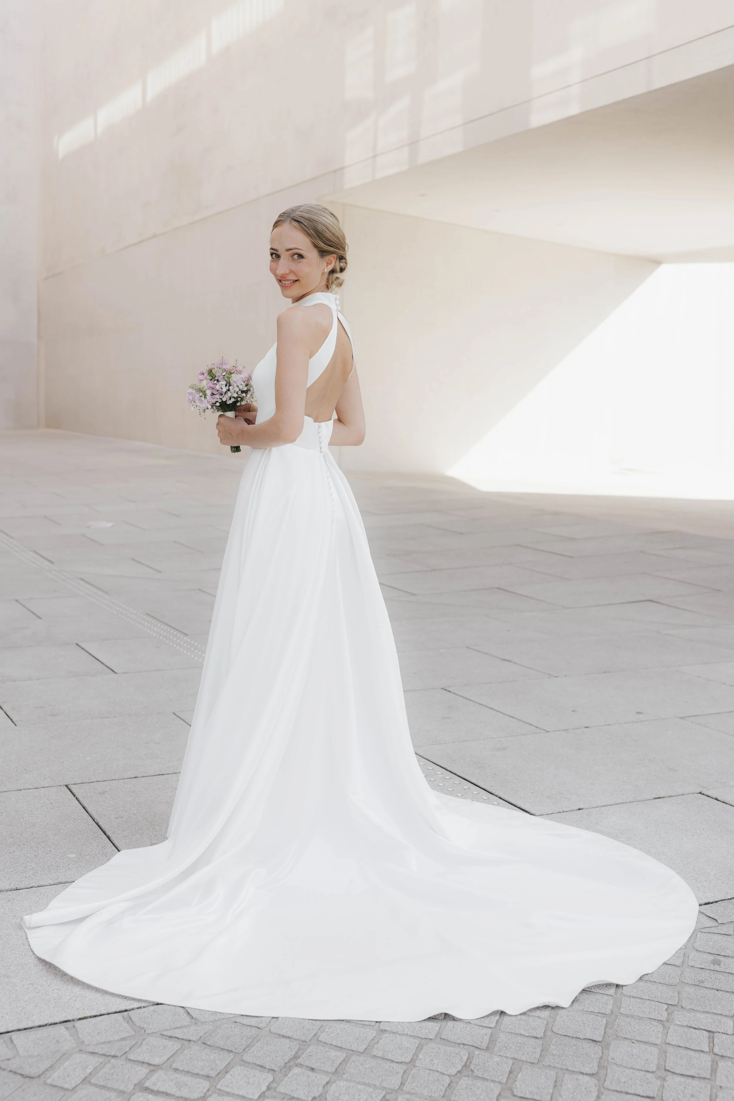 Braut in weißem Hochzeitskleid mit offenem Rücken, hält einen Blumenstrauß, steht auf einer modernen, hellen Außenfläche