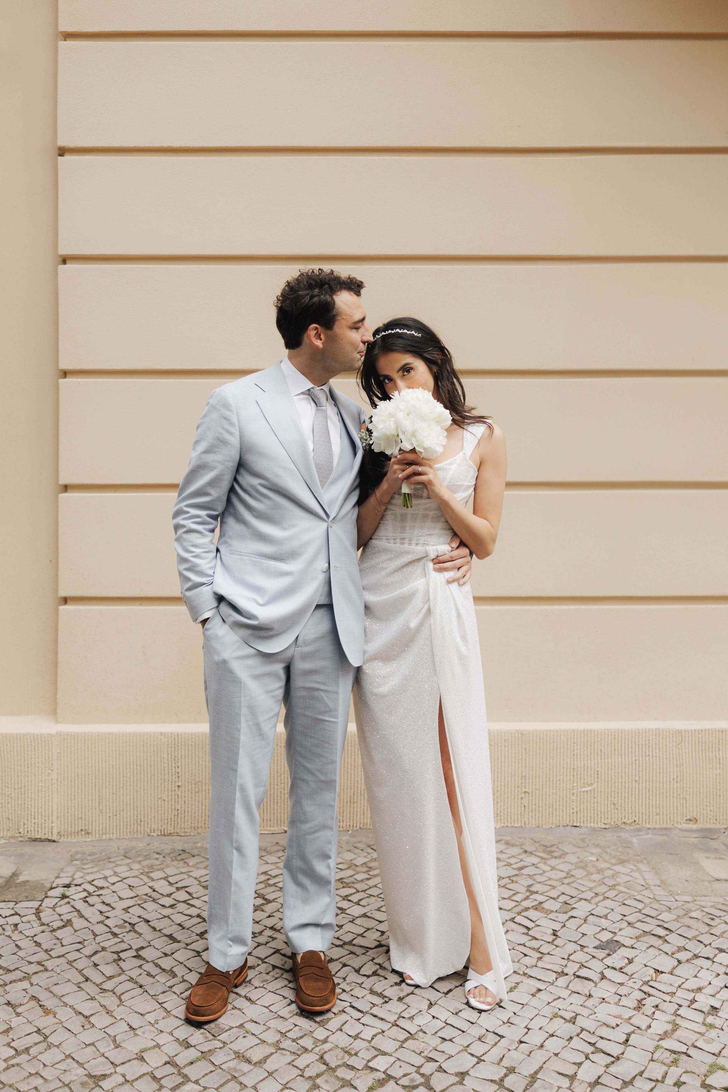 Ein Brautpaar posiert vor einer hellen Wand. Die Braut trägt ein weißes Hochzeitskleid mit einem Schlitz, hält einen Blumenstrauß, und schaut geheimnisvoll. Der Bräutigam trägt einen hellgrauen Anzug und schaut liebevoll auf die Braut.
