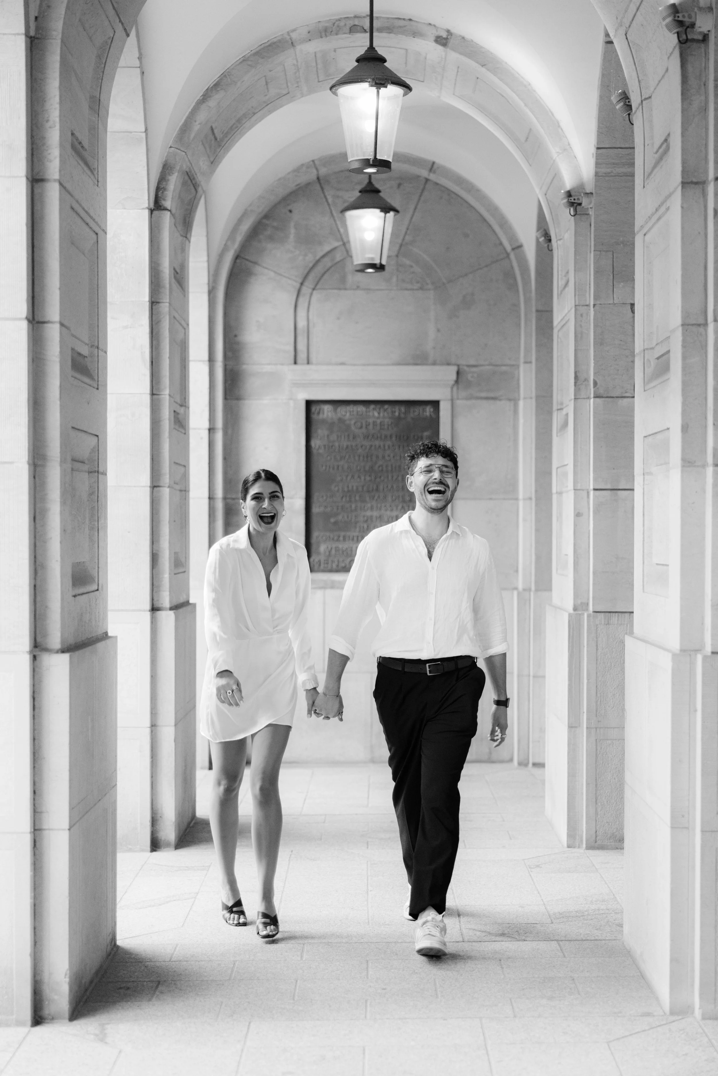 Ein glückliches Paar läuft Hand in Hand durch einen Flur mit Bogen und Deckenlampen, alles in Schwarz-Weiß aufgenommen.