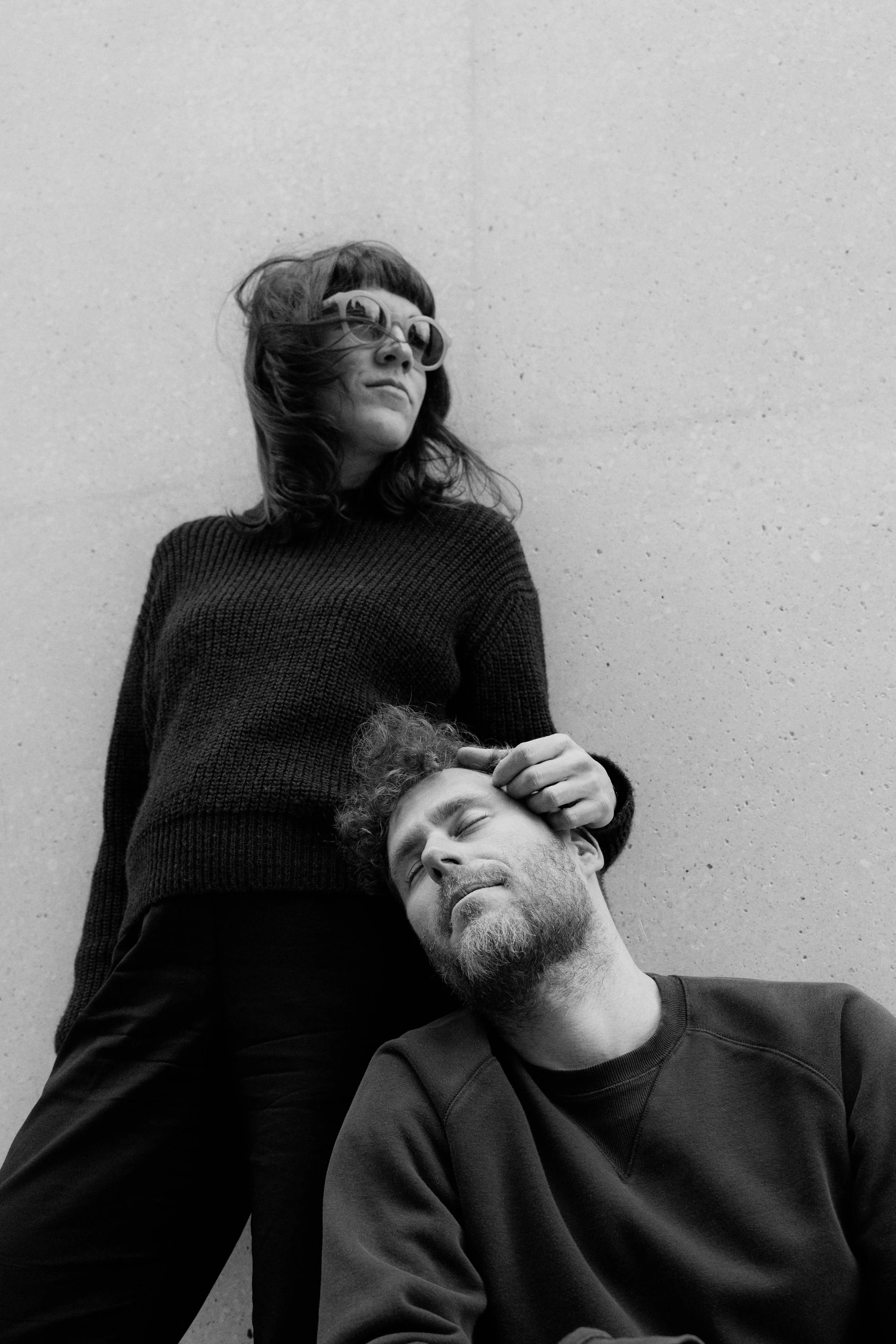 Ein Mann mit Bart sitzt mit geschlossenen Augen, während eine Frau mit dunklem Haar, Sonnenbrille und Pullover ihn berührt, vor einer glatten Wand.