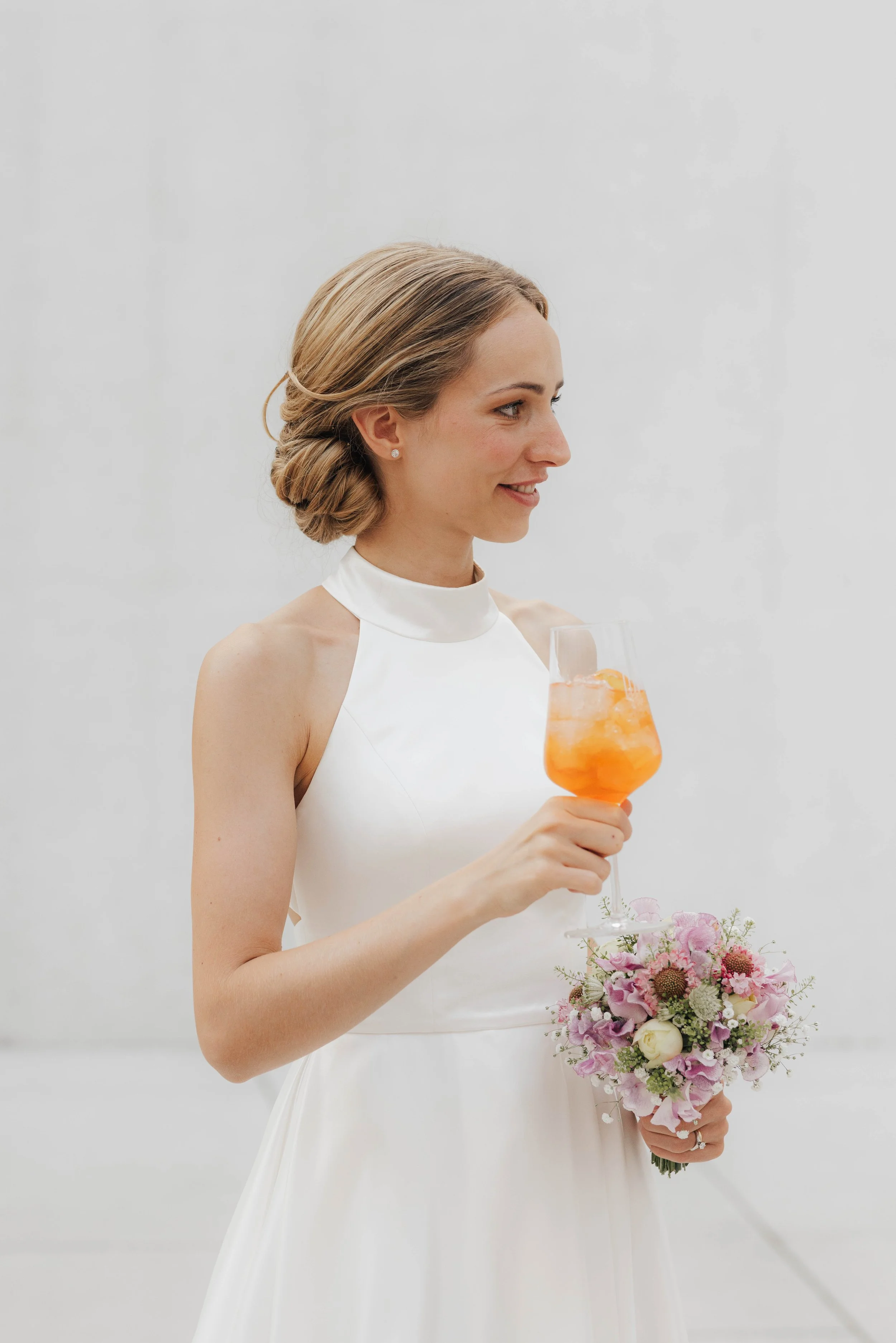 Braut in weißem Kleid hält ein Glas mit orangefarbenem Getränk und Blumenstrauß.