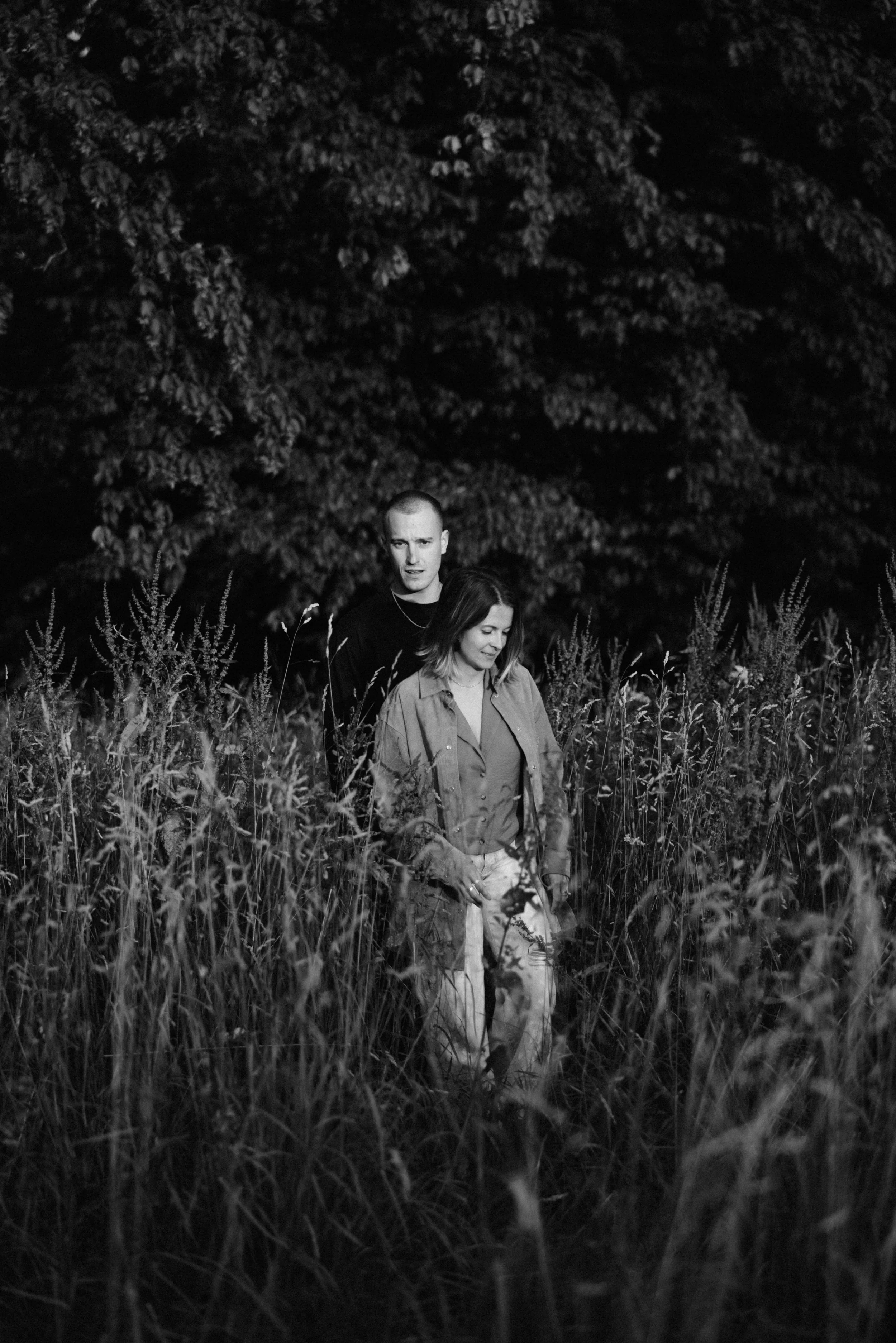 Ein schwarzer und weißer Foto von zwei Menschen, einem Mann und einer Frau, die durch hohes Gras und unter einem Baum bei Nacht gehen.