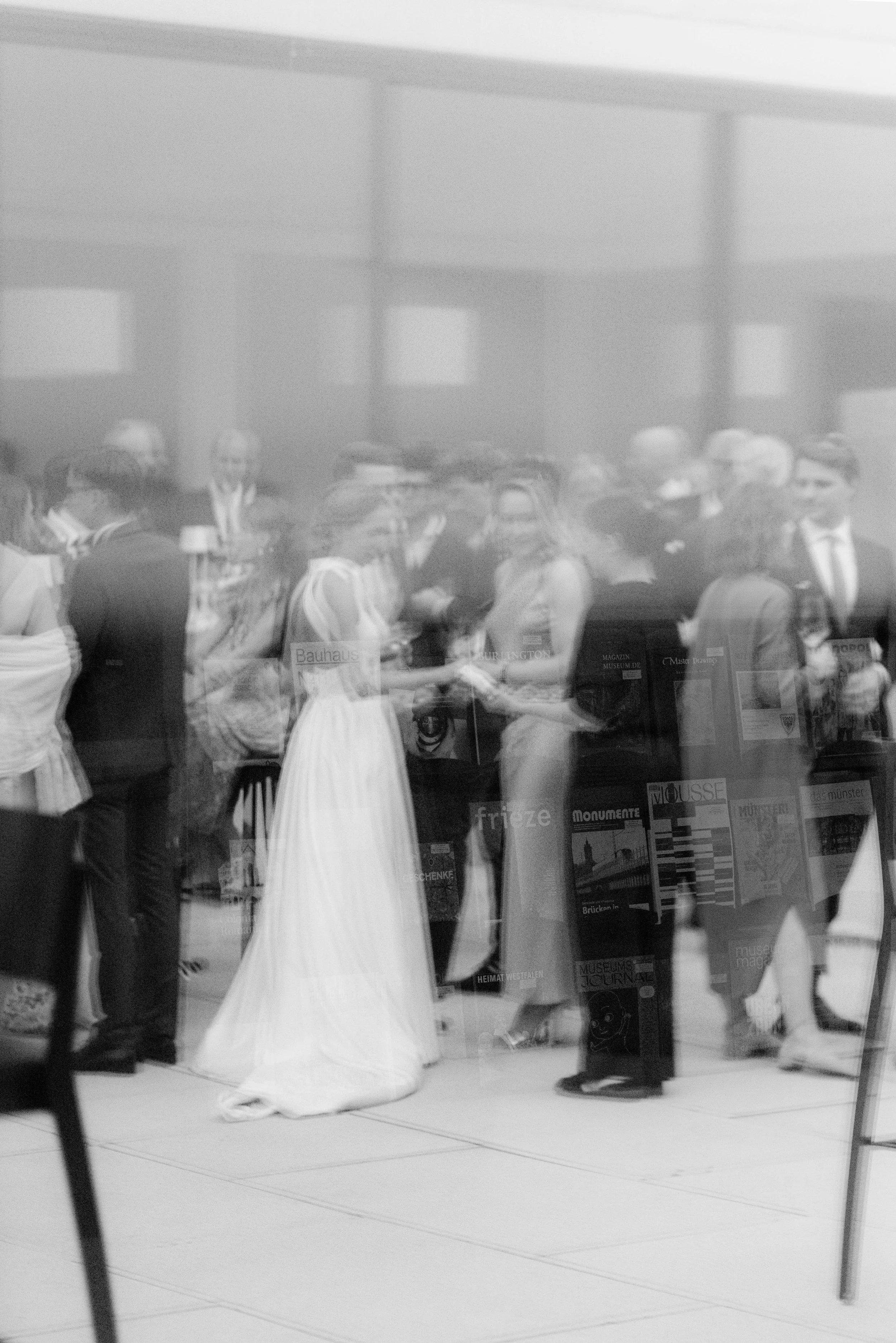 Menschen in formeller Kleidung bei einer Veranstaltung, die durch eine Glaswand reflektiert wird, mit Büchern im Hintergrund.