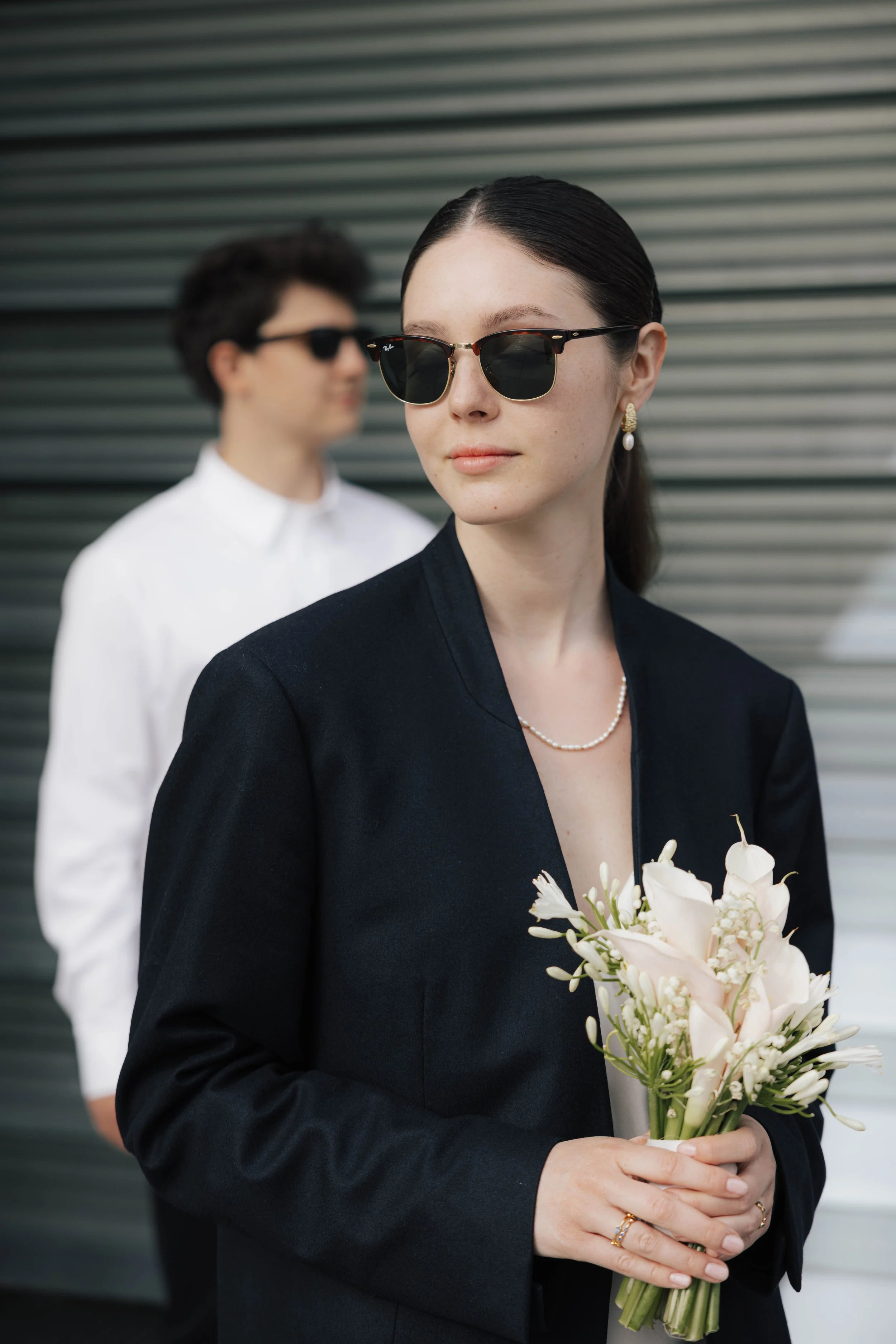 Frau in dunklem Blazer trägt Sonnenbrille, hält einen Blumenstrauß, trägt Perlenkette und Ohrringe, im Hintergrund eine Frau in weißem Hemd und Sonnenbrille vor einer metallischen Wand.