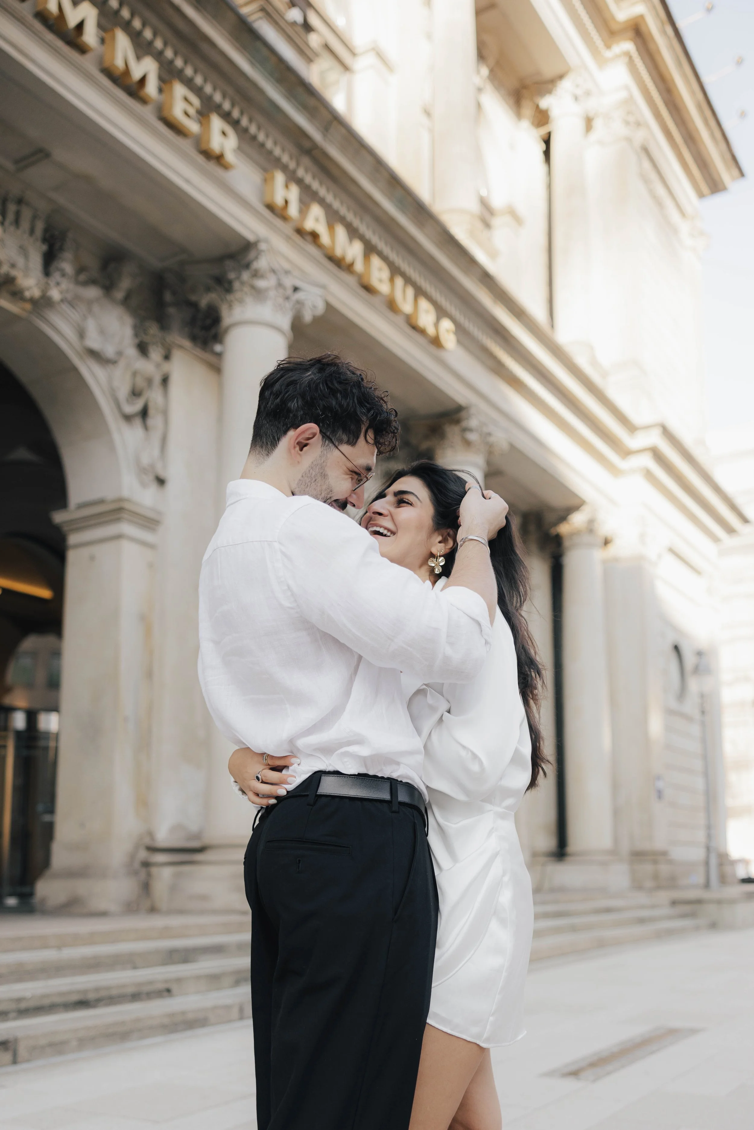 Ein verliebtes Paar umarmt sich vor einem Gebäude, das auf einem Schild "Hamburg" heißt, während sie lachen und sich küssen.