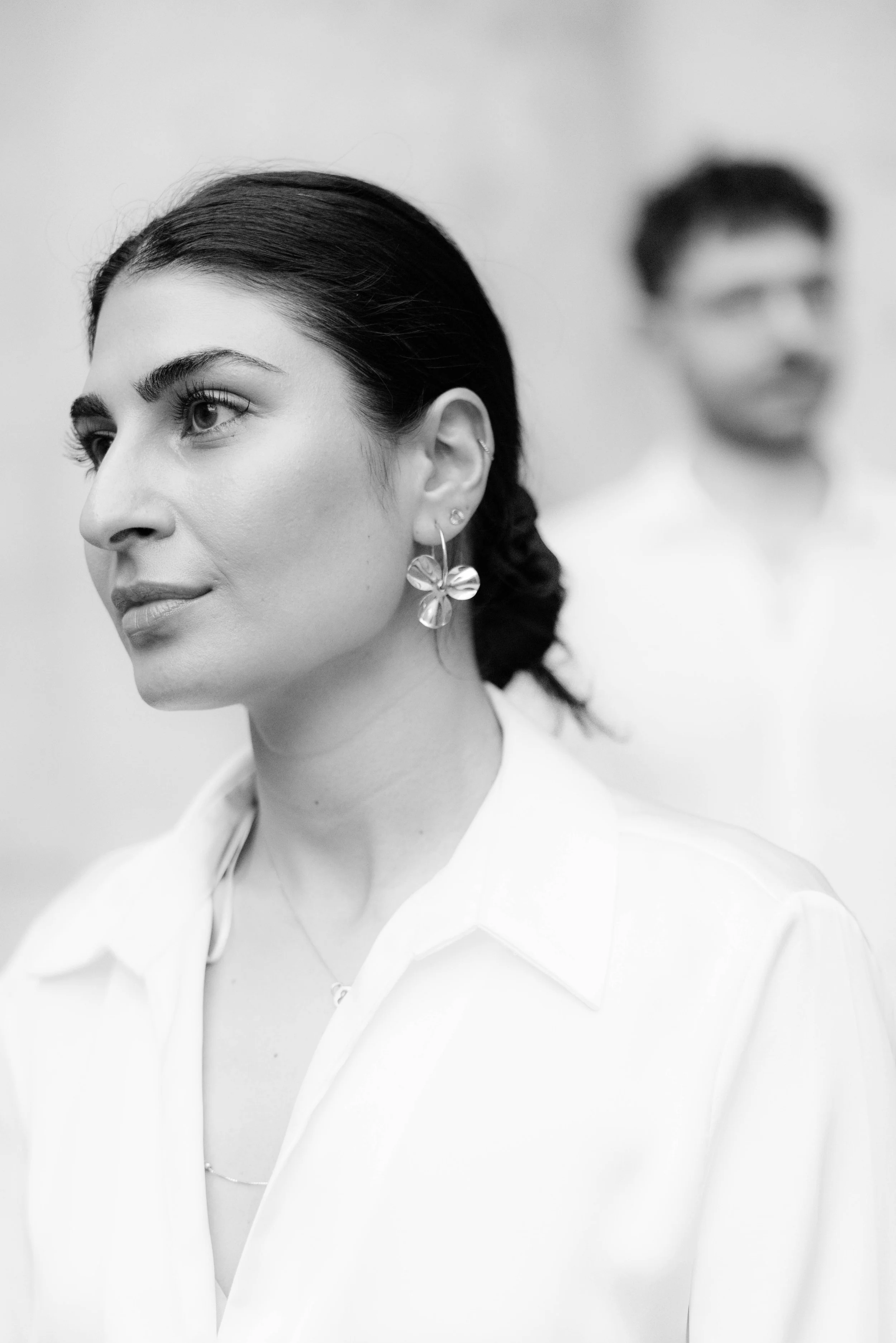 Schwarz-Weiß-Foto einer Frau in Nahaufnahme mit dunklem Haar, großen Ohrringen, hellem Hemd und Make-up, im Hintergrund unscharfer Mann mit Brille.