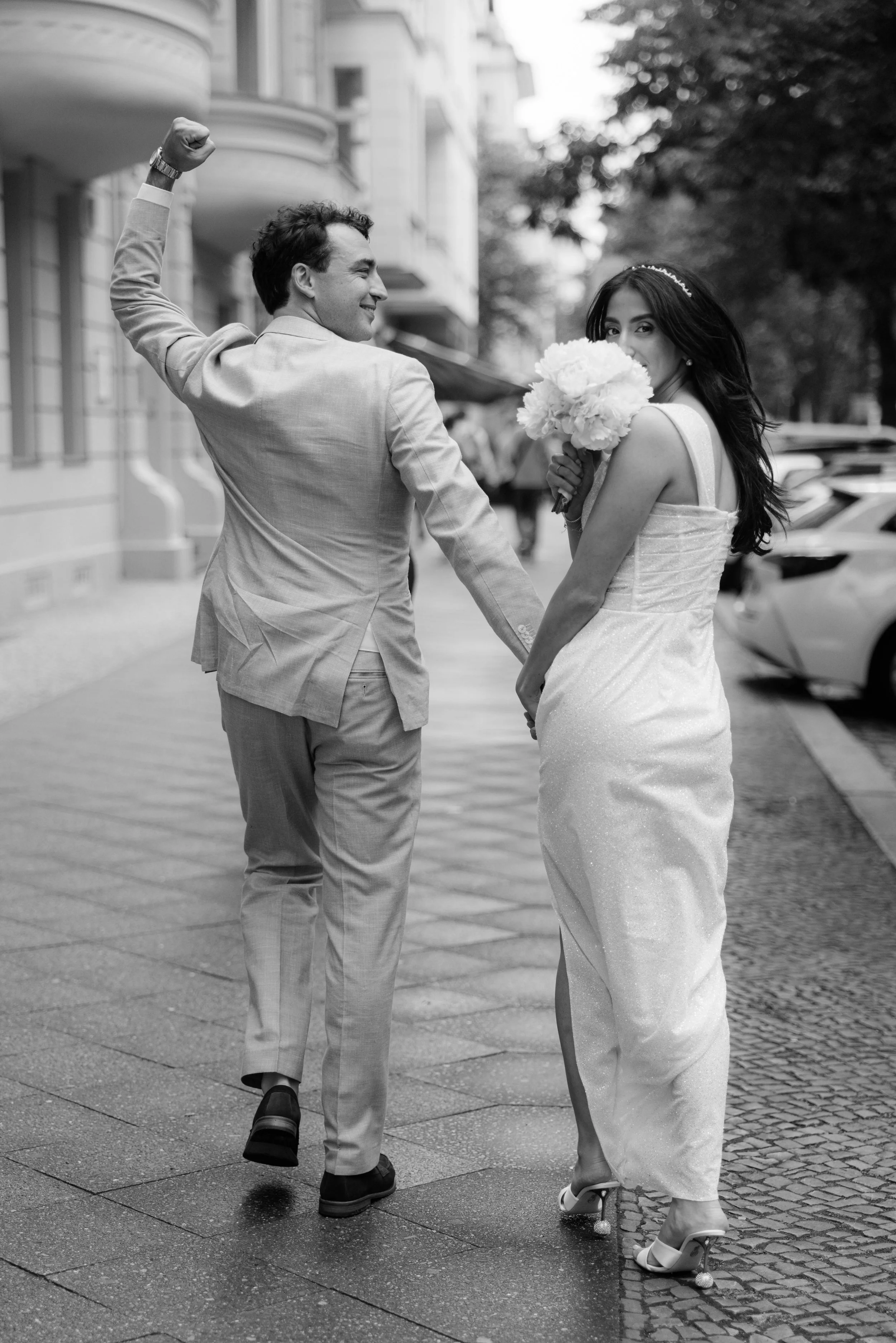 Ein Mann und eine Frau in Hochzeitskleidung gehen auf der Straße, der Mann hebt die Faust in die Luft, die Frau hält einen Blumenstrauß und schaut über die Schulter, beide lachen.