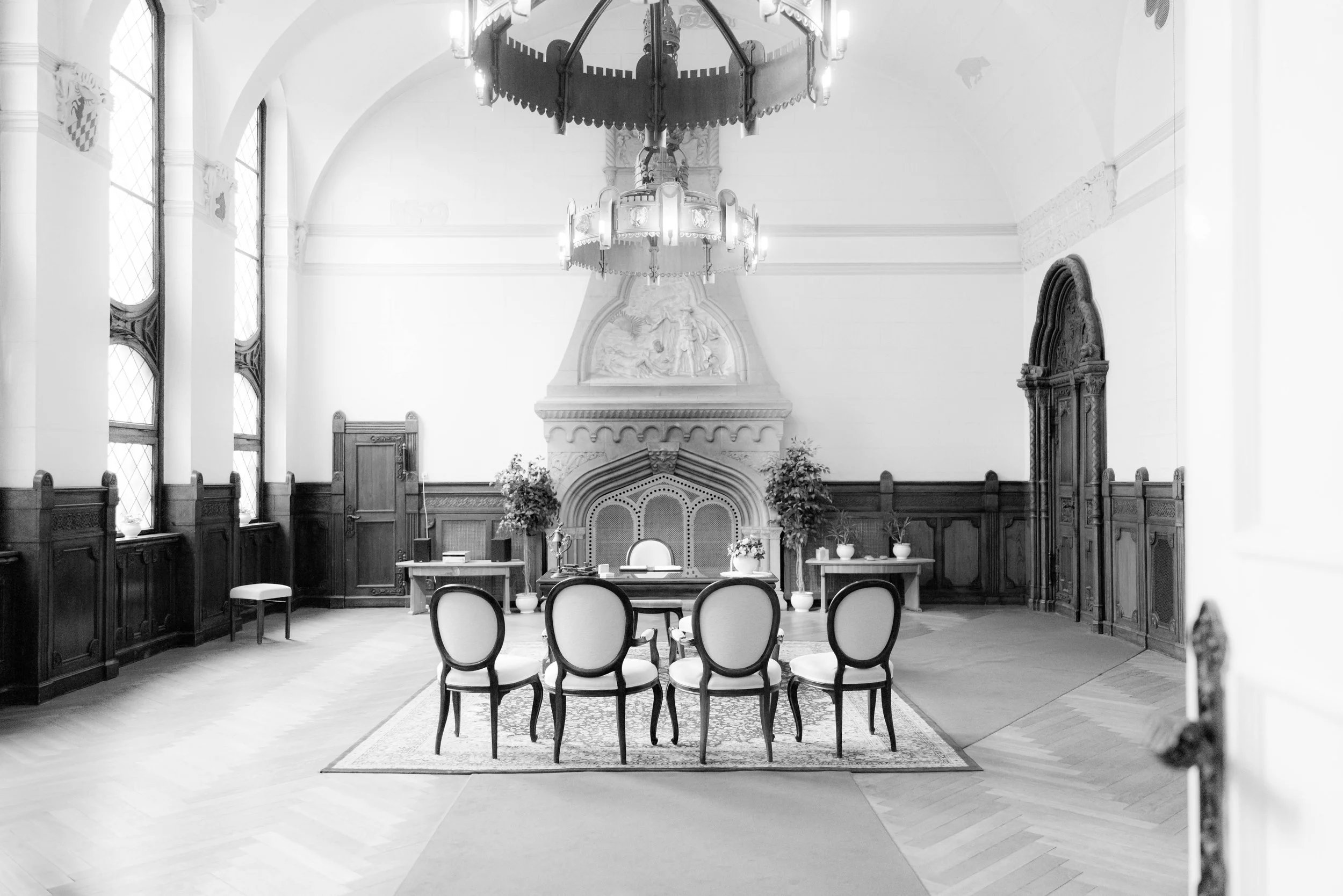 Ein historischer, nachdenklicher Saal mit einem großen Kamin, mehreren Stühlen, einem schweren Holztisch, Pflanzen und einem Kronleuchter.