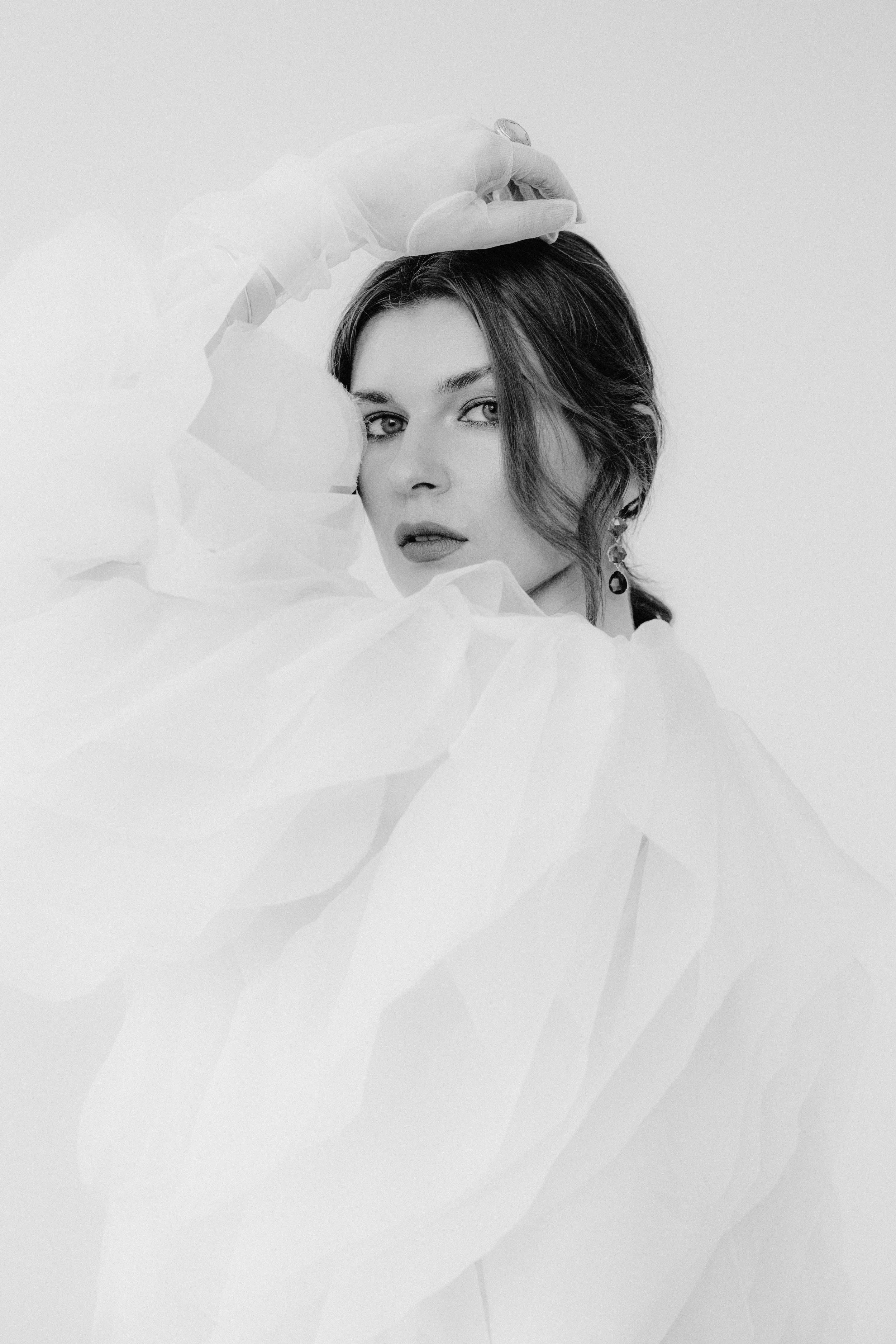 Schwarz-weiß-Fotografie eines Mädchens mit dunklen Haaren, die in eine Richtung blickt, umgeben von weichen, transparent wirkenden Stoffen, die eine dramatische und elegante Atmosphäre schaffen.